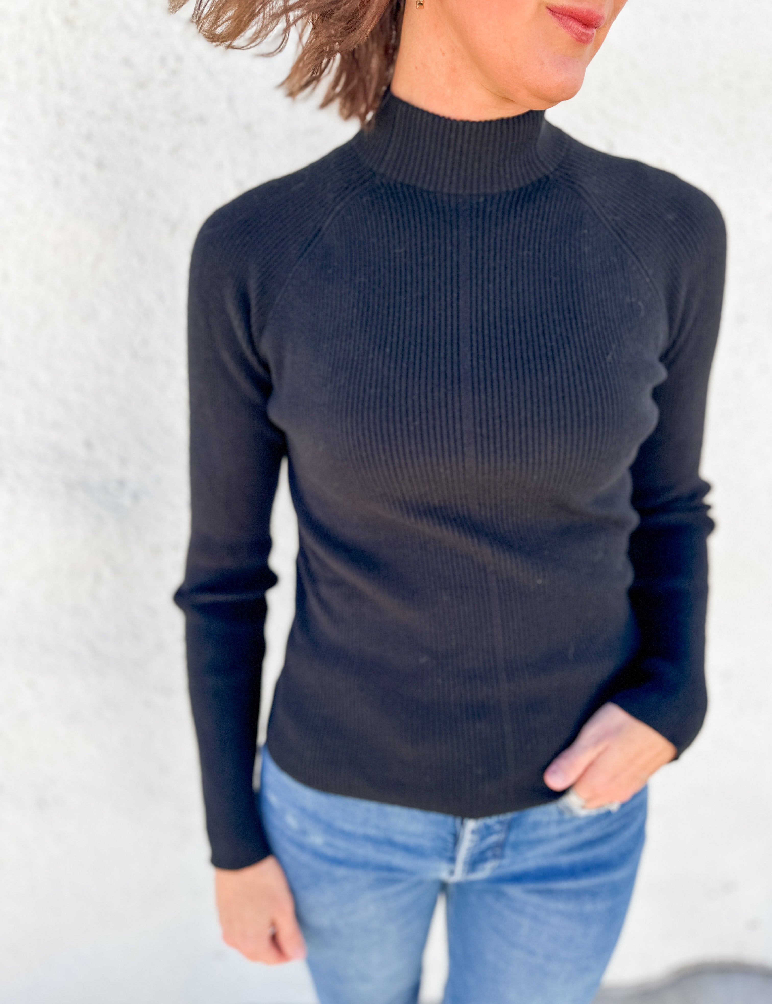 A woman wearing a black ribbed mock neck long-sleeve sweater and light blue jeans, standing against a textured white wall.