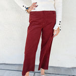 A person wearing burgundy cropped pants with a small white polka dot pattern, styled with a cream ribbed knit sweater and beige ballet flats against a white stucco wall.