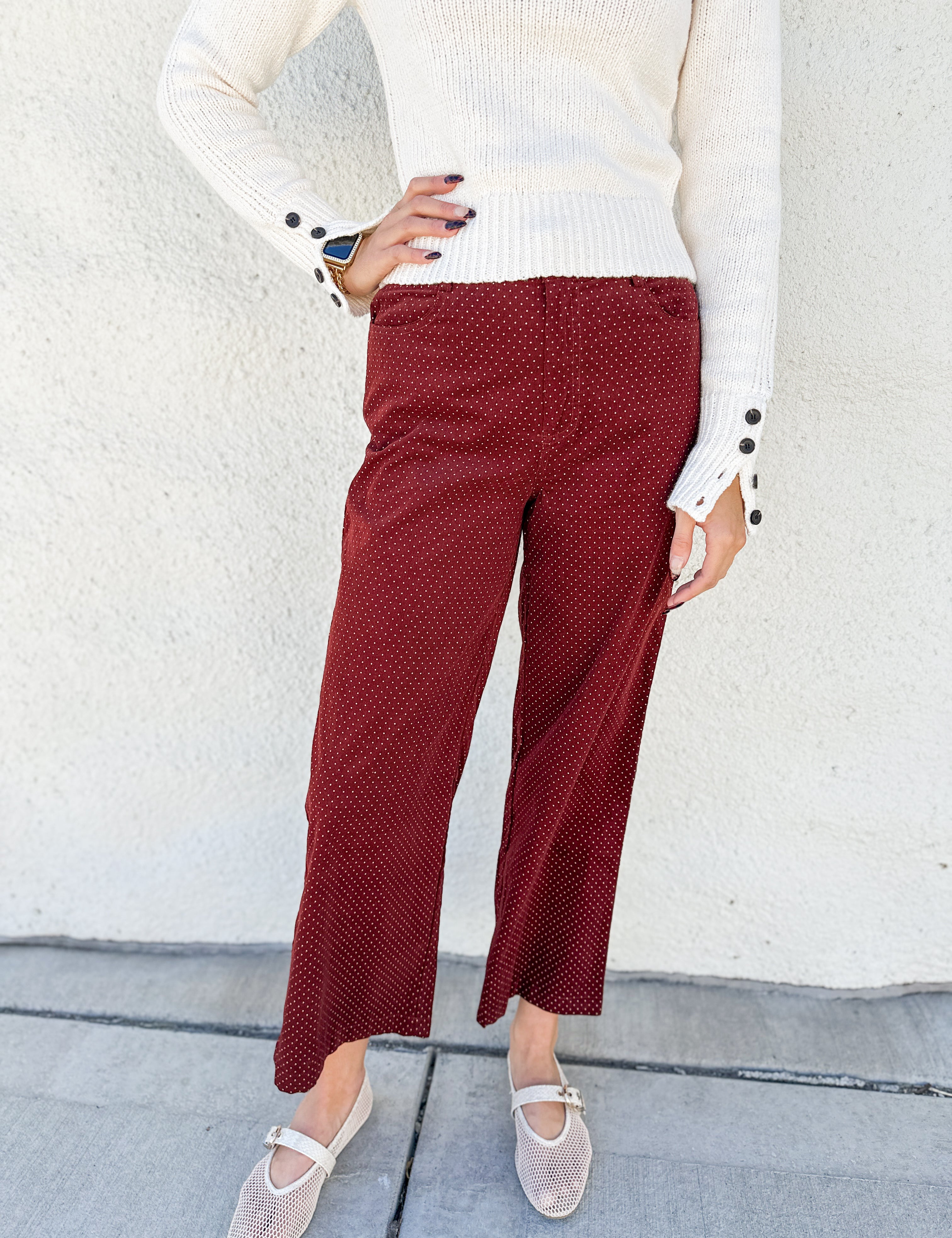 A person wearing burgundy cropped pants with a small white polka dot pattern, styled with a cream ribbed knit sweater and beige ballet flats against a white stucco wall.