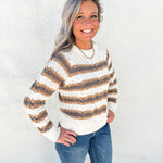 A smiling woman with gray hair stands against a white textured wall wearing a cream knit sweater with brown and tan horizontal stripes, blue jeans, and a gold necklace.