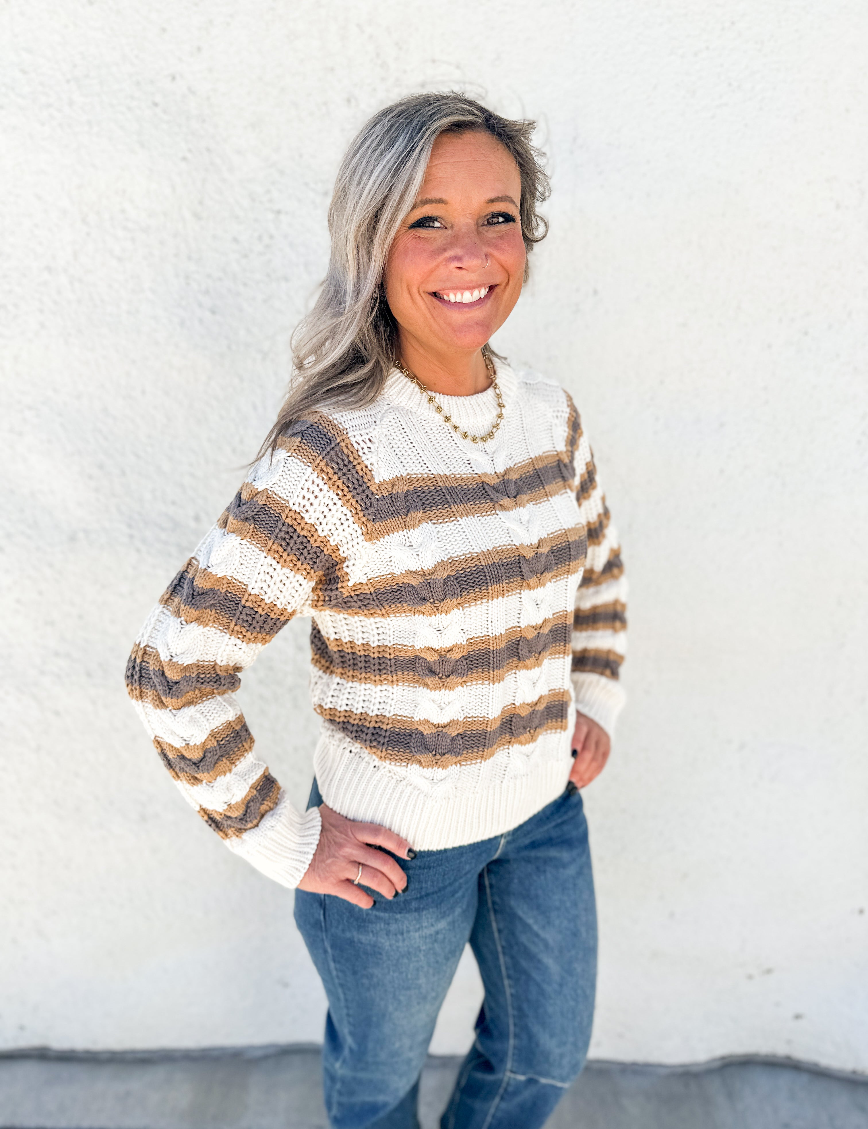 A smiling woman with gray hair stands against a white textured wall wearing a cream knit sweater with brown and tan horizontal stripes, blue jeans, and a gold necklace.