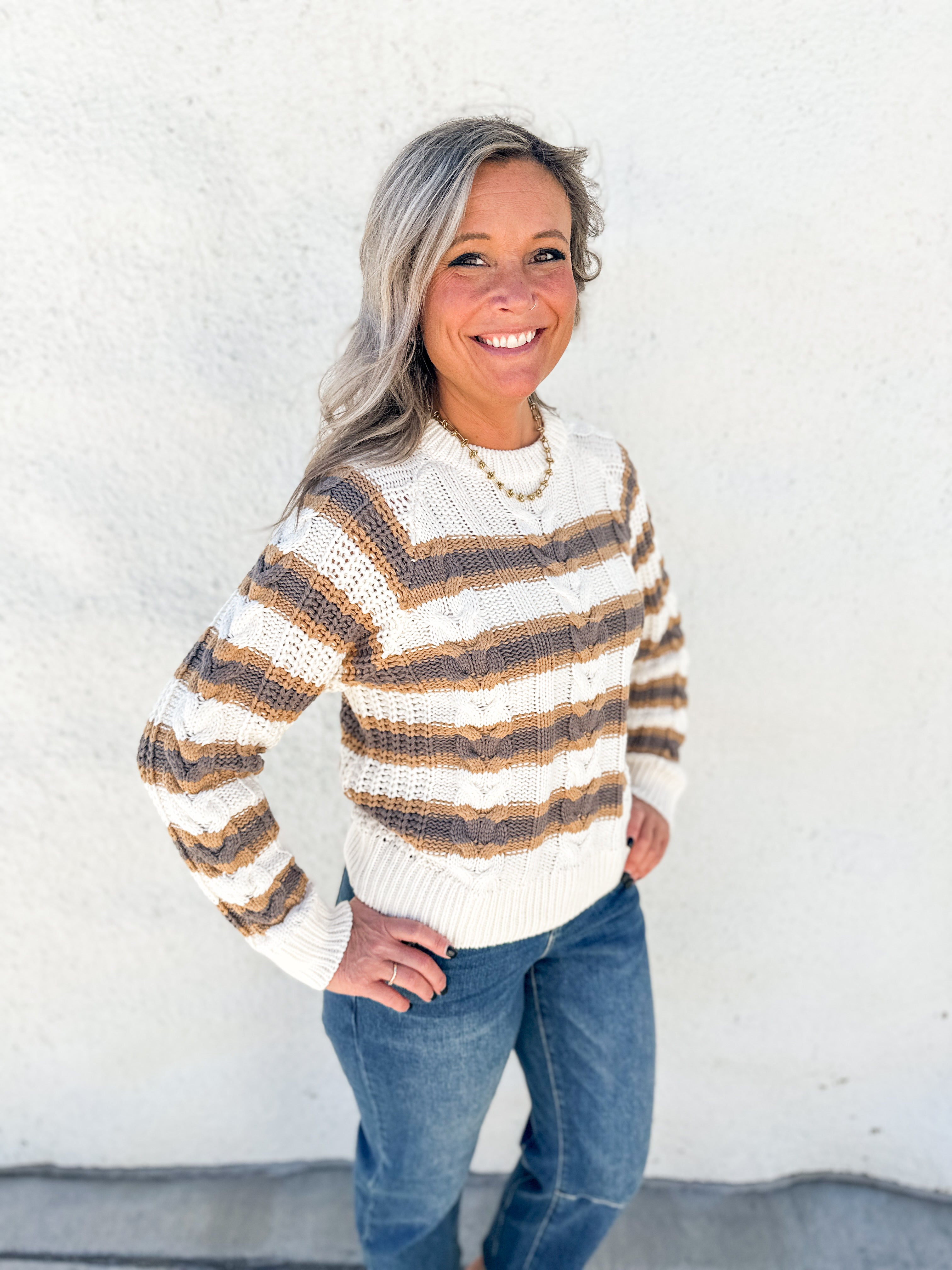 A smiling woman with gray hair stands against a white textured wall wearing a cream knit sweater with brown and tan horizontal stripes, blue jeans, and a gold necklace.