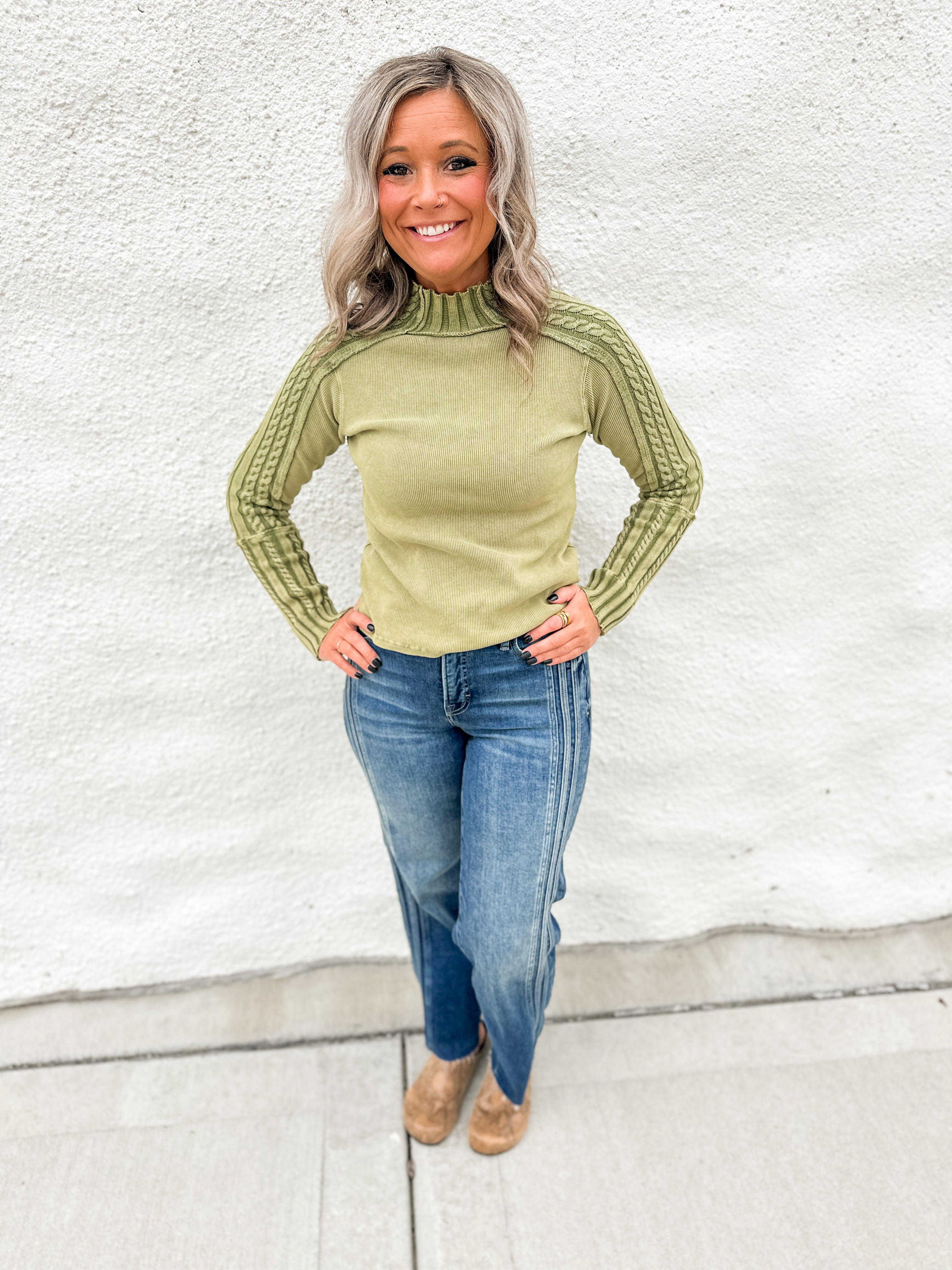 A woman wearing a mixed media green thermal top with cable knit sleeves and wearing blue denim jeans with seaming down the sides and snake print mule clogs in front of a white wall.