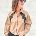 A woman wearing a tan long-sleeve button-up shirt with black lace applique on the chest, paired with black pants and black sunglasses, standing against a white textured wall.