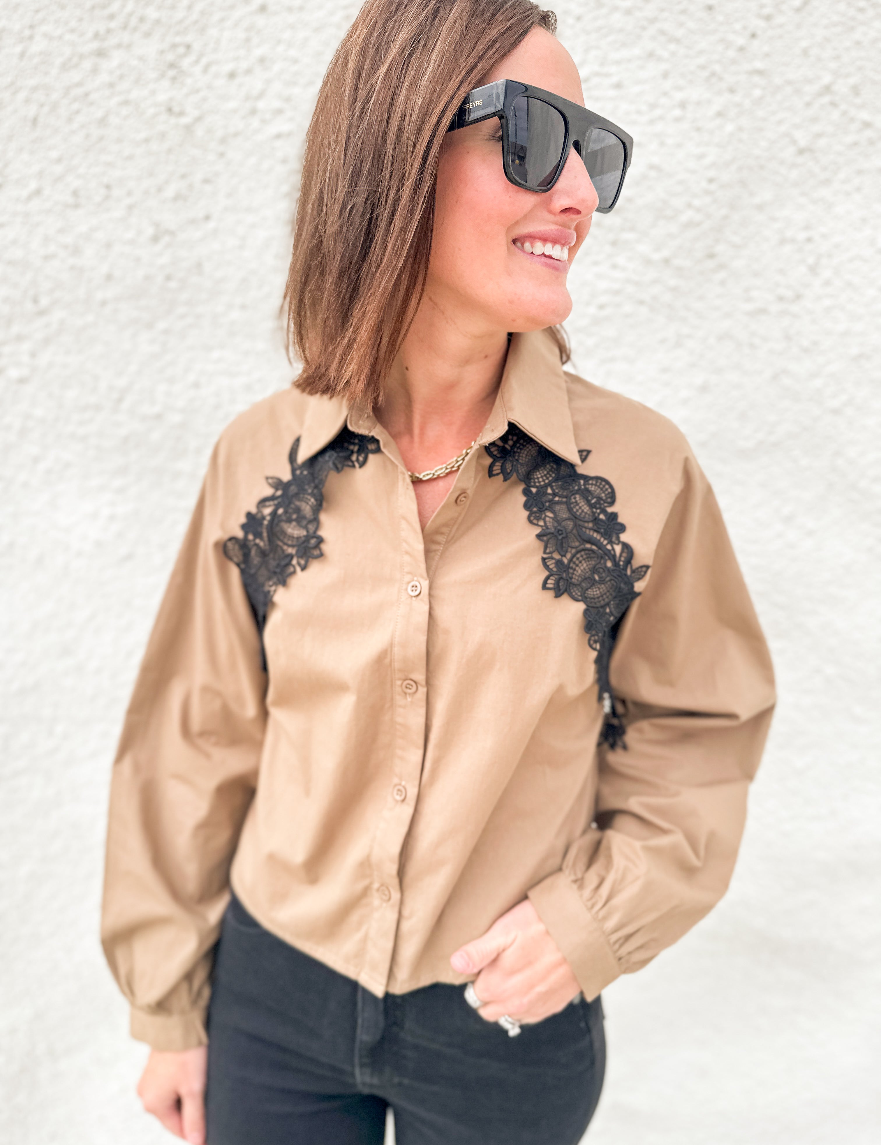 A woman wearing a tan long-sleeve button-up shirt with black lace applique on the chest, paired with black pants and black sunglasses, standing against a white textured wall.