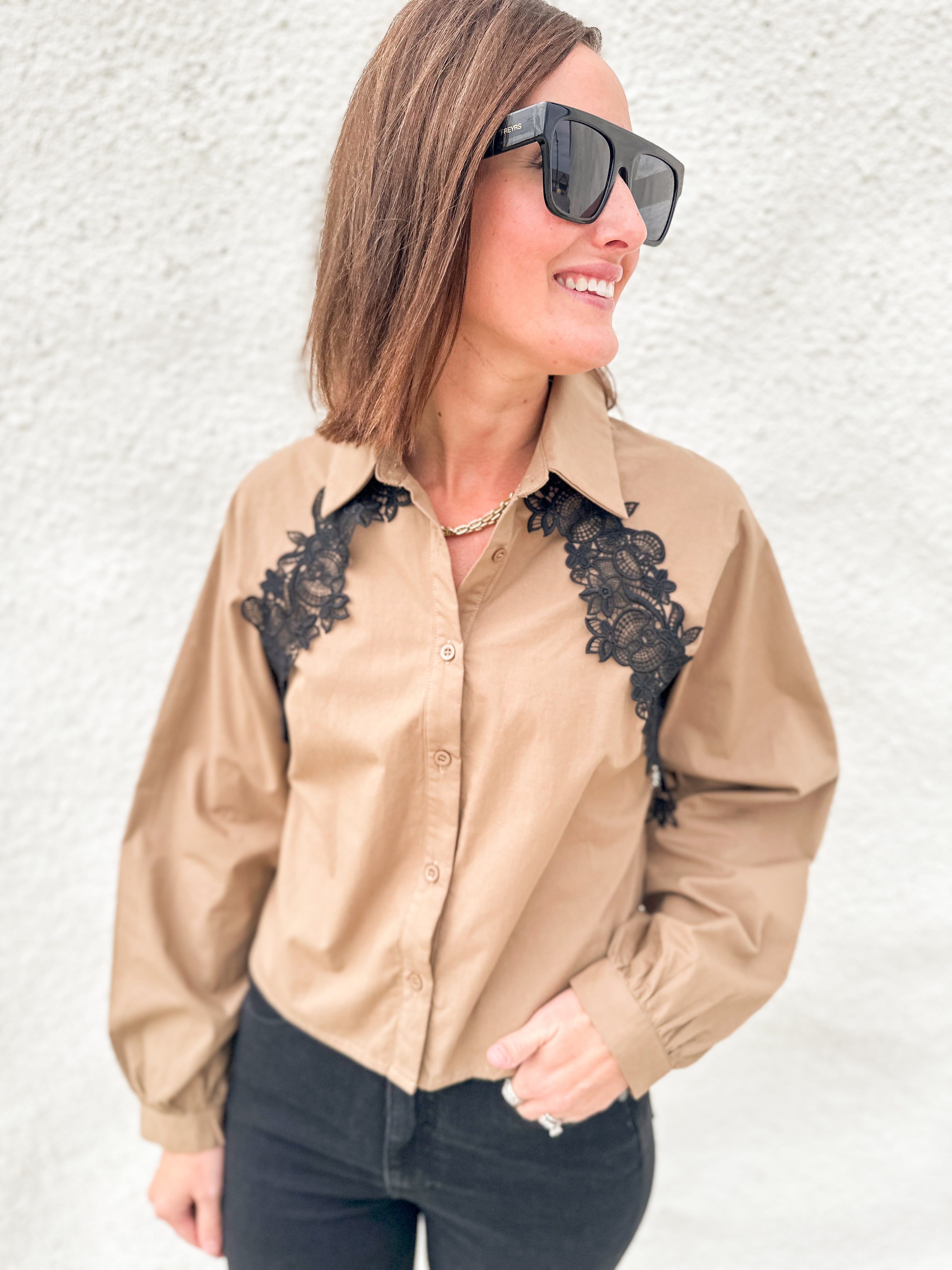 A woman wearing a tan long-sleeve button-up shirt with black lace applique on the chest, paired with black pants and black sunglasses, standing against a white textured wall.