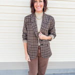 A woman stands in front of a cream-colored siding wall wearing a brown and beige plaid blazer by Dear John Denim over a beige turtleneck, paired with brown pants.