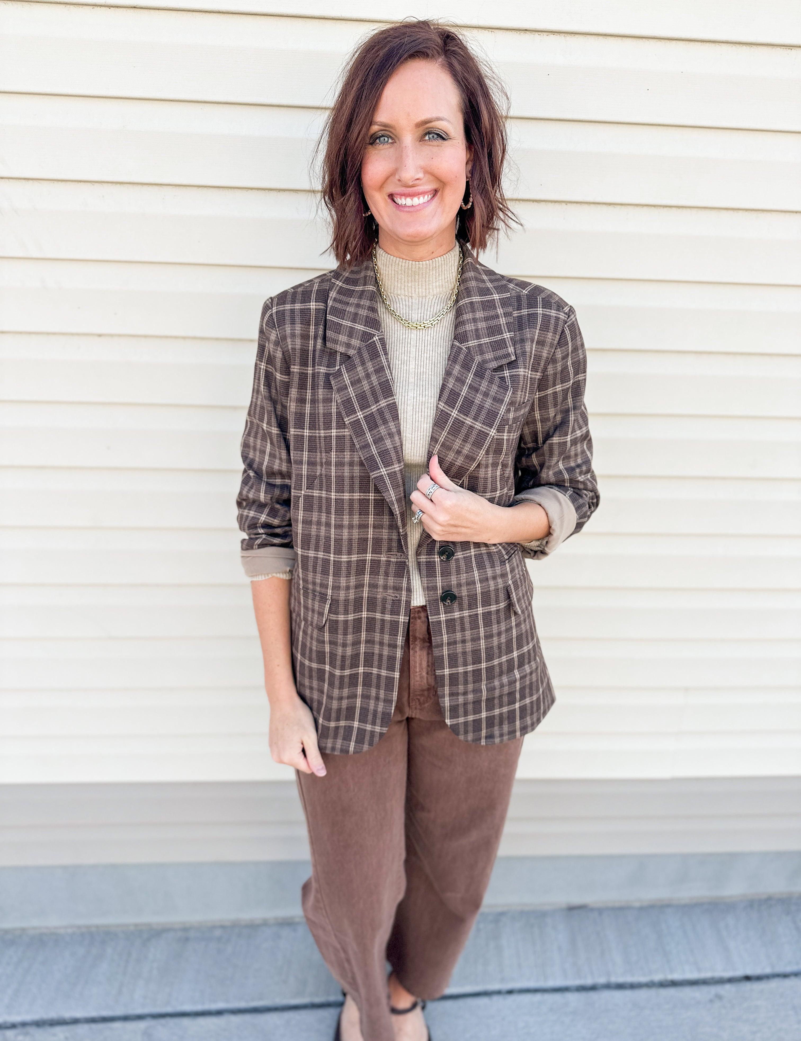 A woman stands in front of a cream-colored siding wall wearing a brown and beige plaid blazer by Dear John Denim over a beige turtleneck, paired with brown pants.