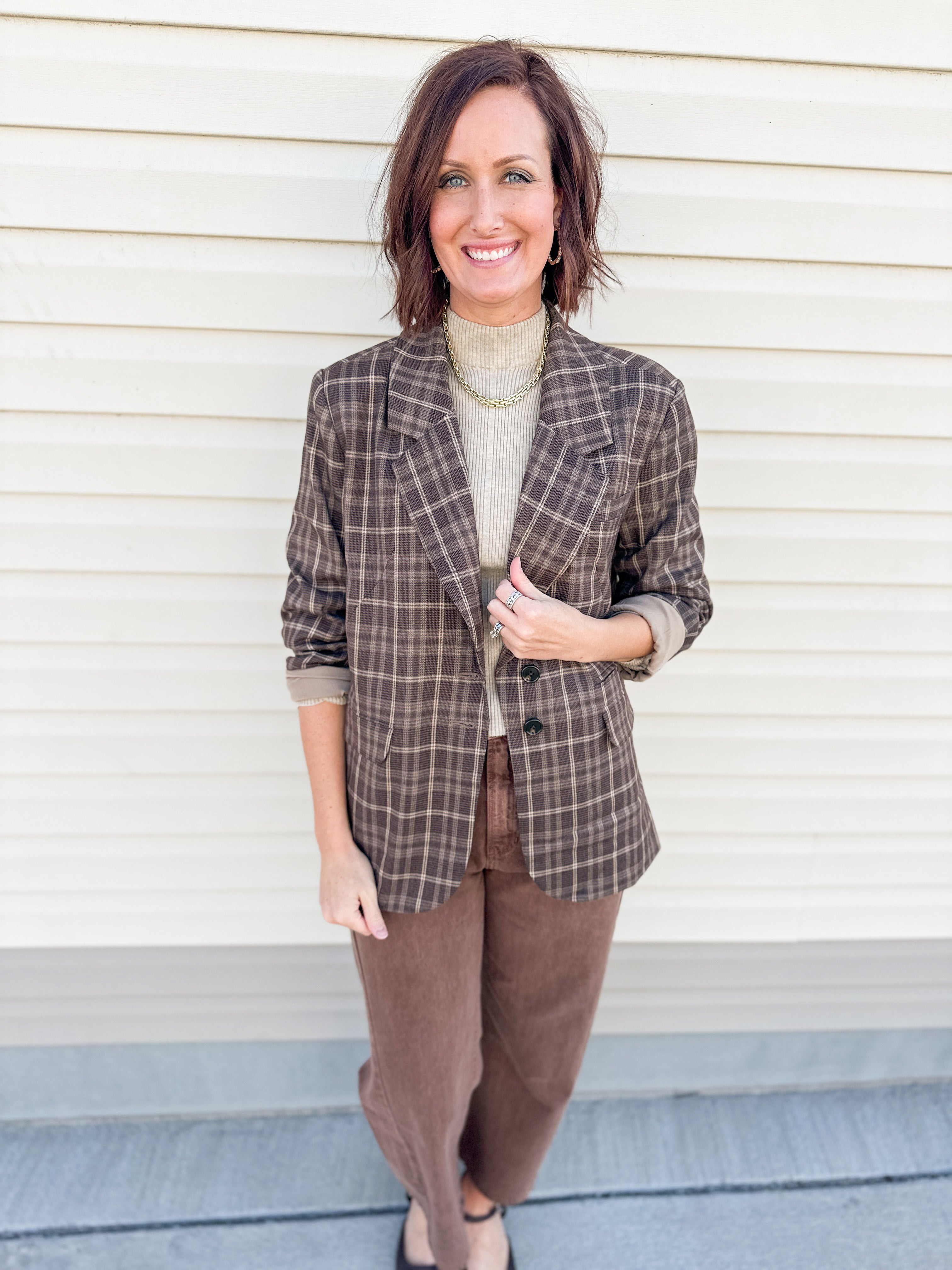A woman stands in front of a cream-colored siding wall wearing a brown and beige plaid blazer by Dear John Denim over a beige turtleneck, paired with brown pants.