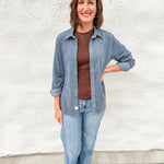 A woman wearing a chocolate brown tank layered with a light blue corduroy shirt jacket wearing light blue denim against a white wall.