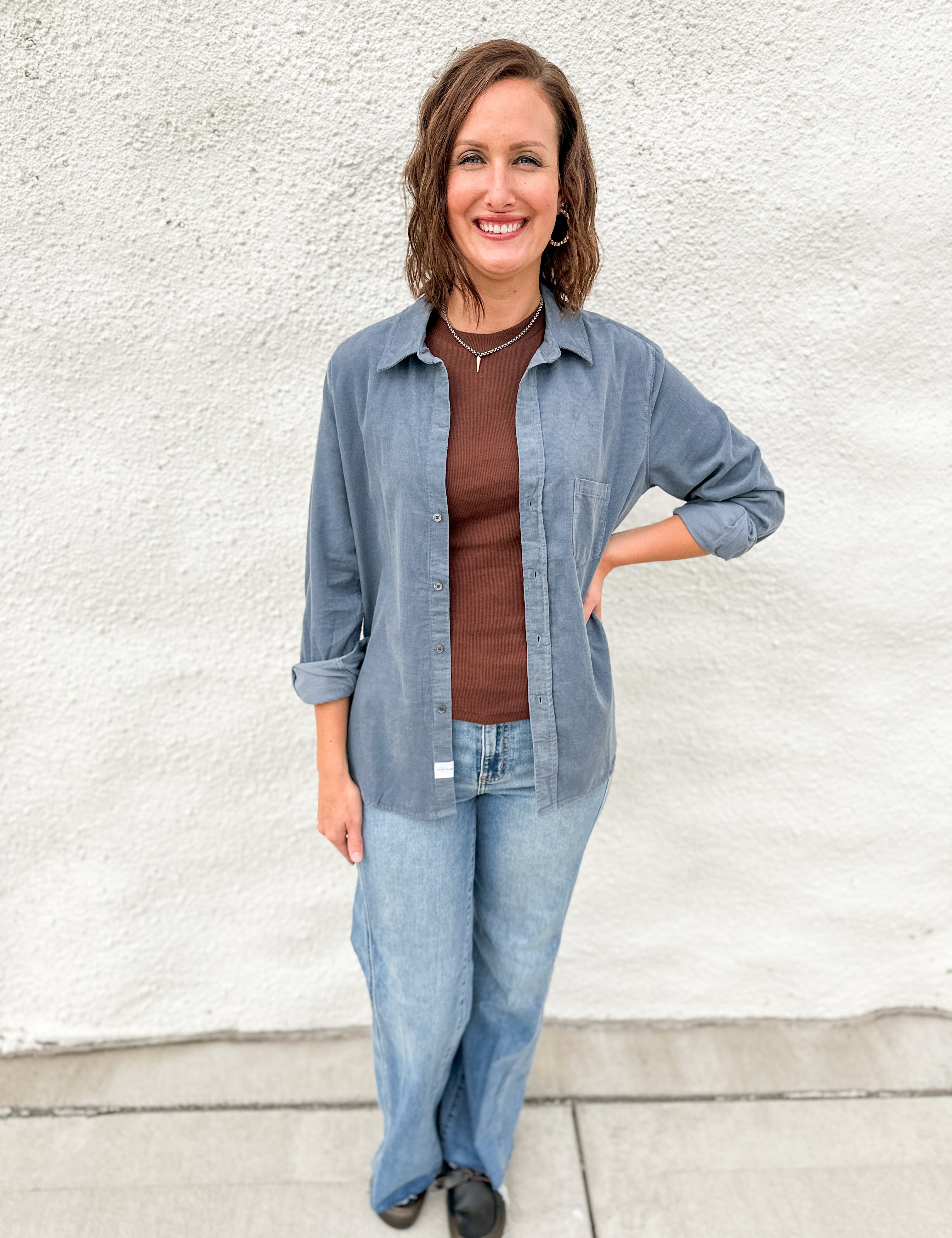 A woman wearing a chocolate brown tank layered with a light blue corduroy shirt jacket wearing light blue denim against a white wall.