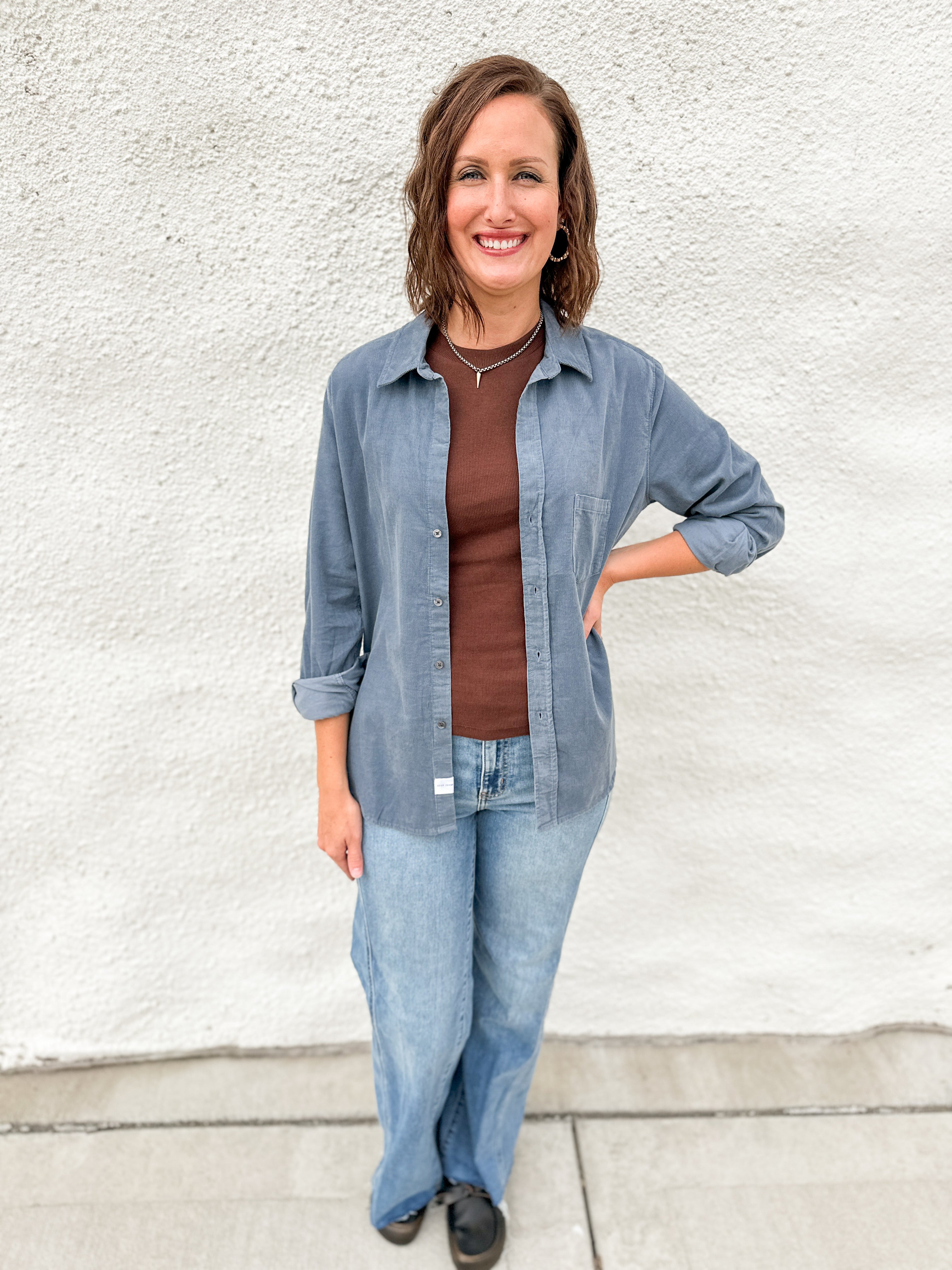 A woman wearing a chocolate brown tank layered with a light blue corduroy shirt jacket wearing light blue denim against a white wall.