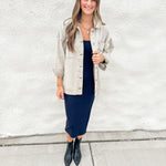 A woman stands against a white textured wall wearing a light grey/green oversized denim jacket over a navy midi dress, paired with black pointed ankle boots.