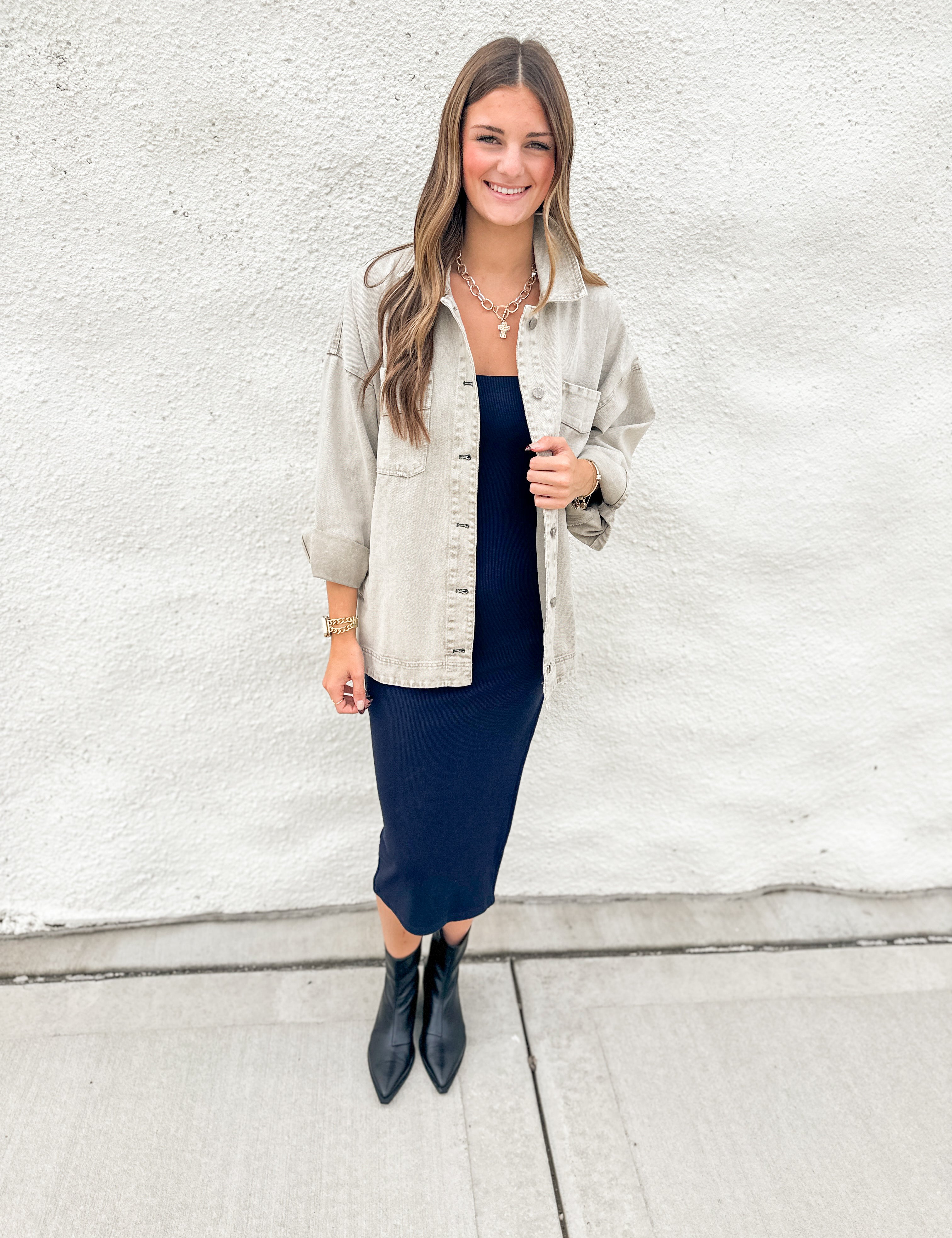A woman stands against a white textured wall wearing a light grey/green oversized denim jacket over a navy midi dress, paired with black pointed ankle boots.