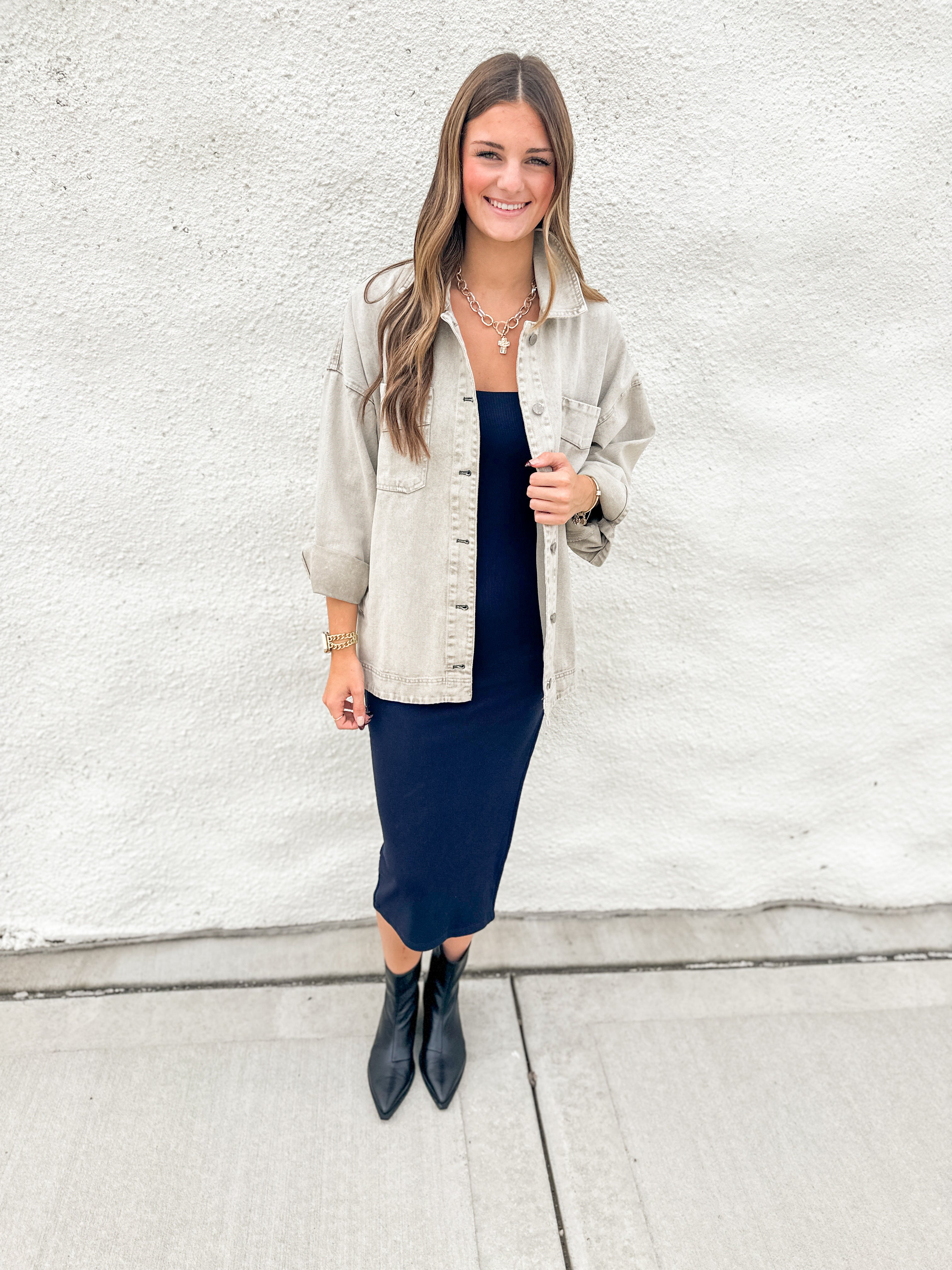 A woman stands against a white textured wall wearing a light grey/green oversized denim jacket over a navy midi dress, paired with black pointed ankle boots.