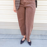 A person wearing brown cropped wide-leg corduroy pants, styled with a plaid blazer and dark pointed flats, standing against a beige siding background.