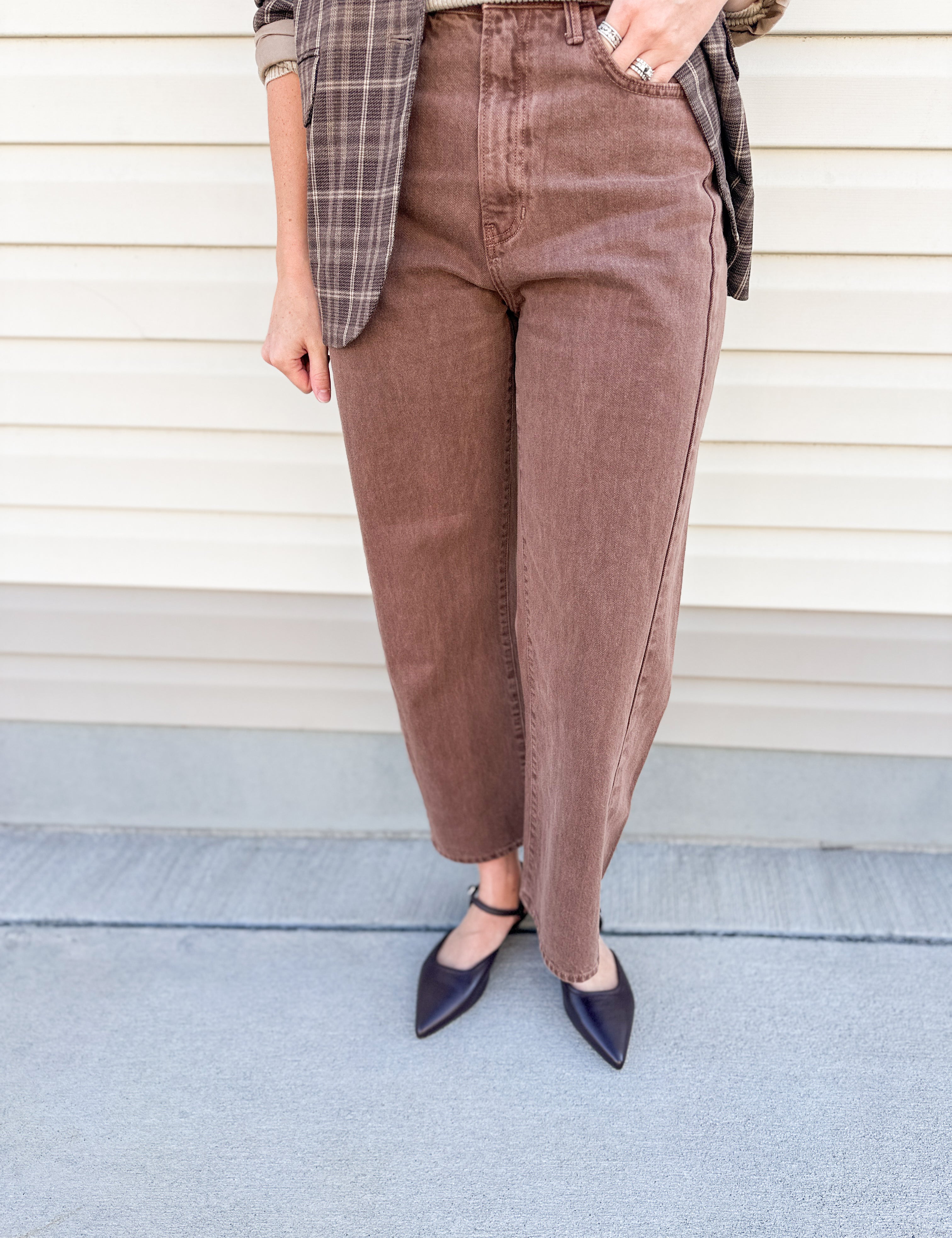 A person wearing brown cropped wide-leg corduroy pants, styled with a plaid blazer and dark pointed flats, standing against a beige siding background.