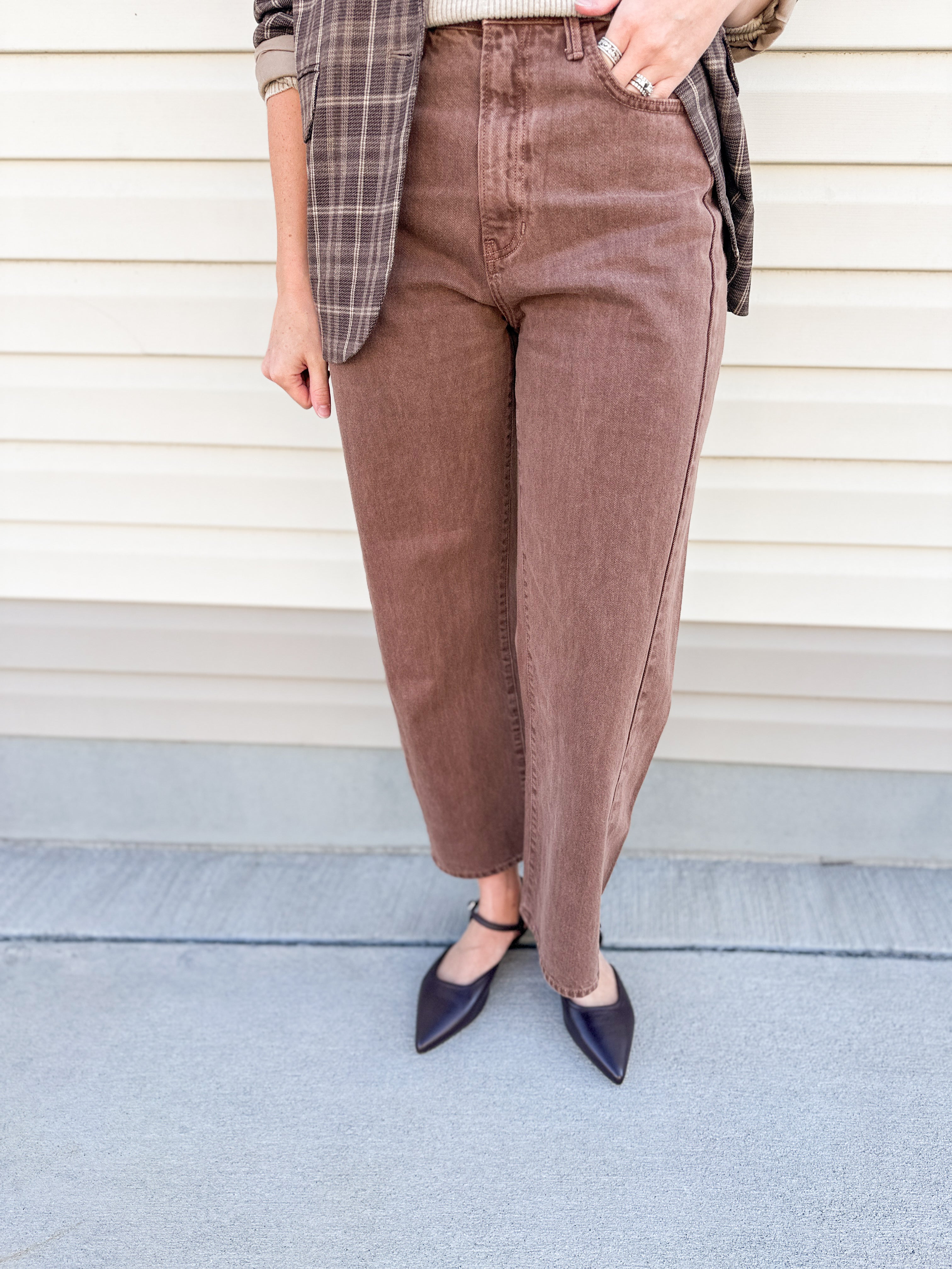 A person wearing brown cropped wide-leg corduroy pants, styled with a plaid blazer and dark pointed flats, standing against a beige siding background.