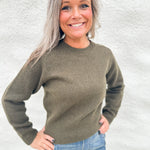 A smiling woman with gray hair wearing an olive green wool knit crewneck sweater and blue jeans, standing against a light gray textured wall.