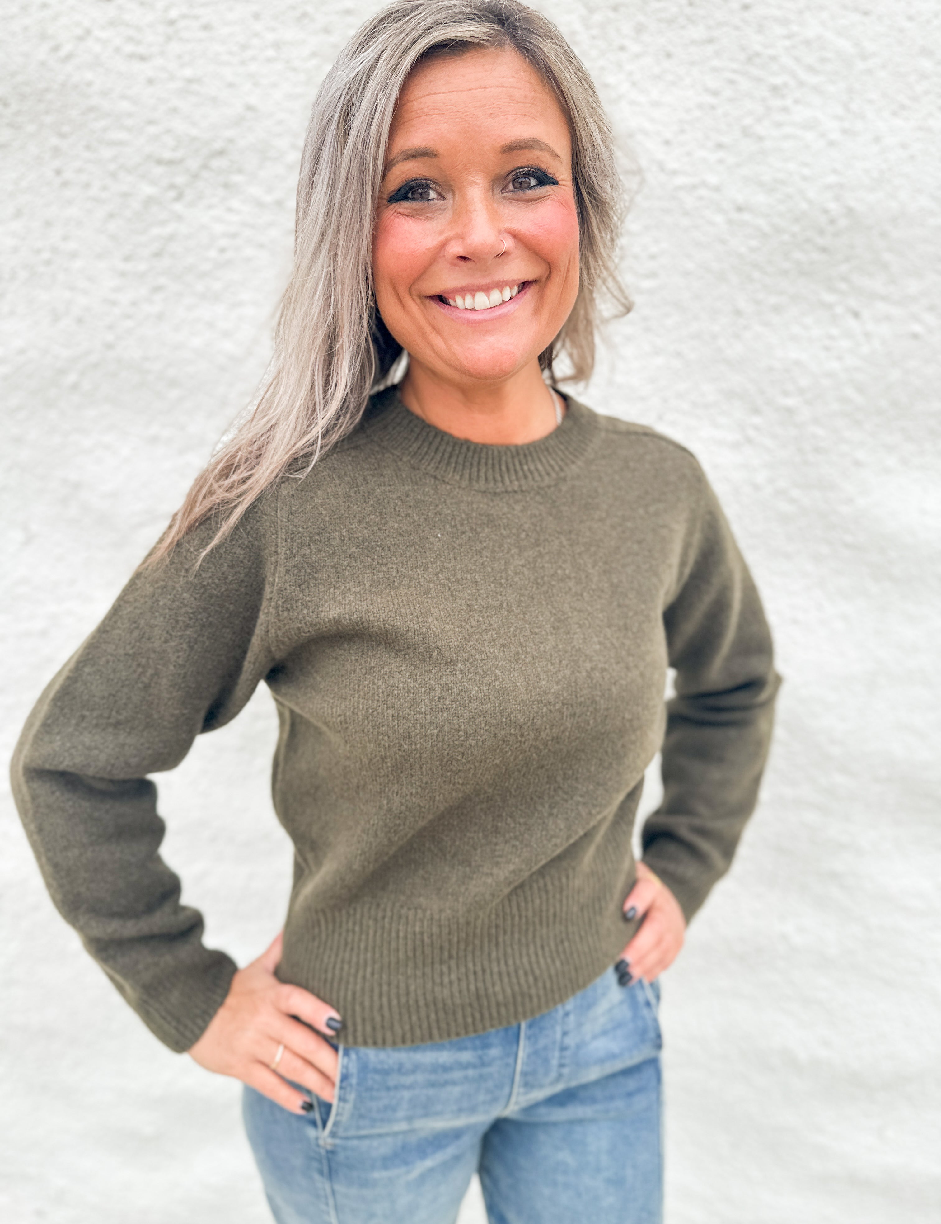 A smiling woman with gray hair wearing an olive green wool knit crewneck sweater and blue jeans, standing against a light gray textured wall.