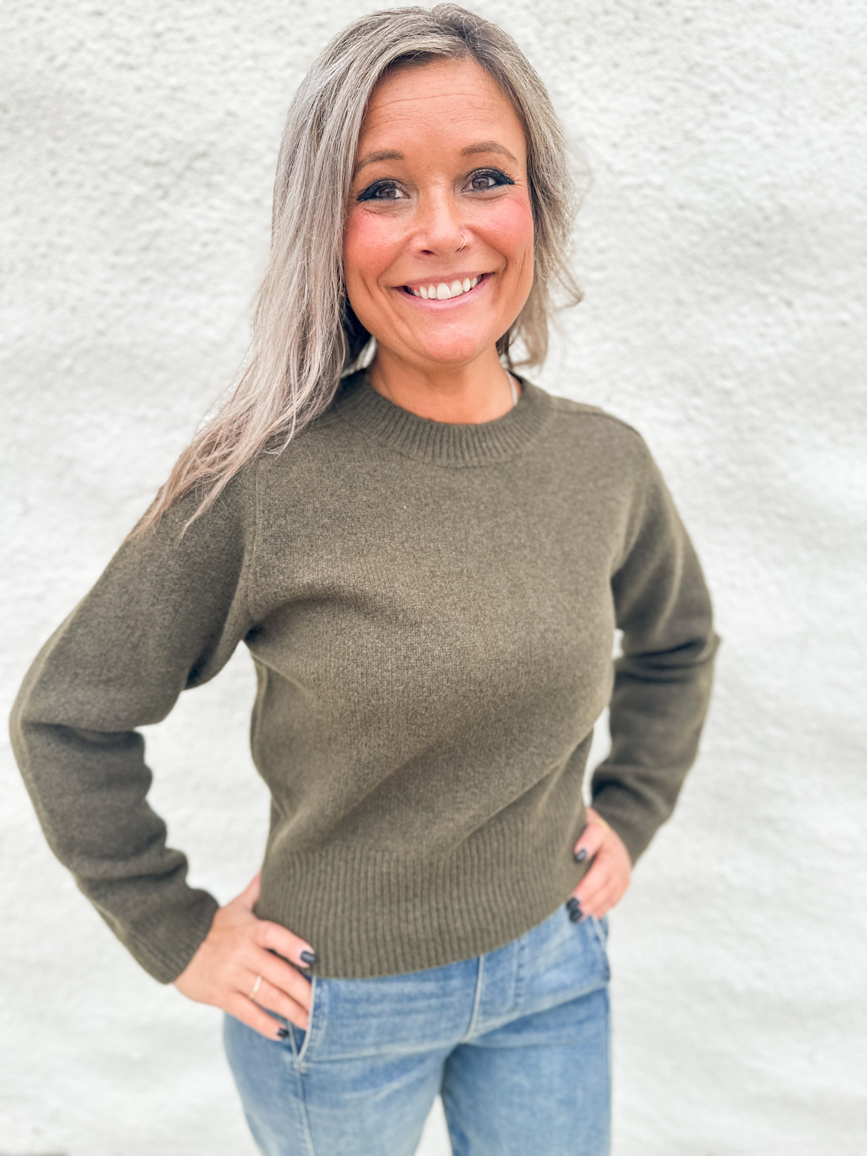 A smiling woman with gray hair wearing an olive green wool knit crewneck sweater and blue jeans, standing against a light gray textured wall.