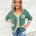 A girl wearing a green and white striped cardigan with GB on the front pocket area wearing blue jeans in front of a white wall.