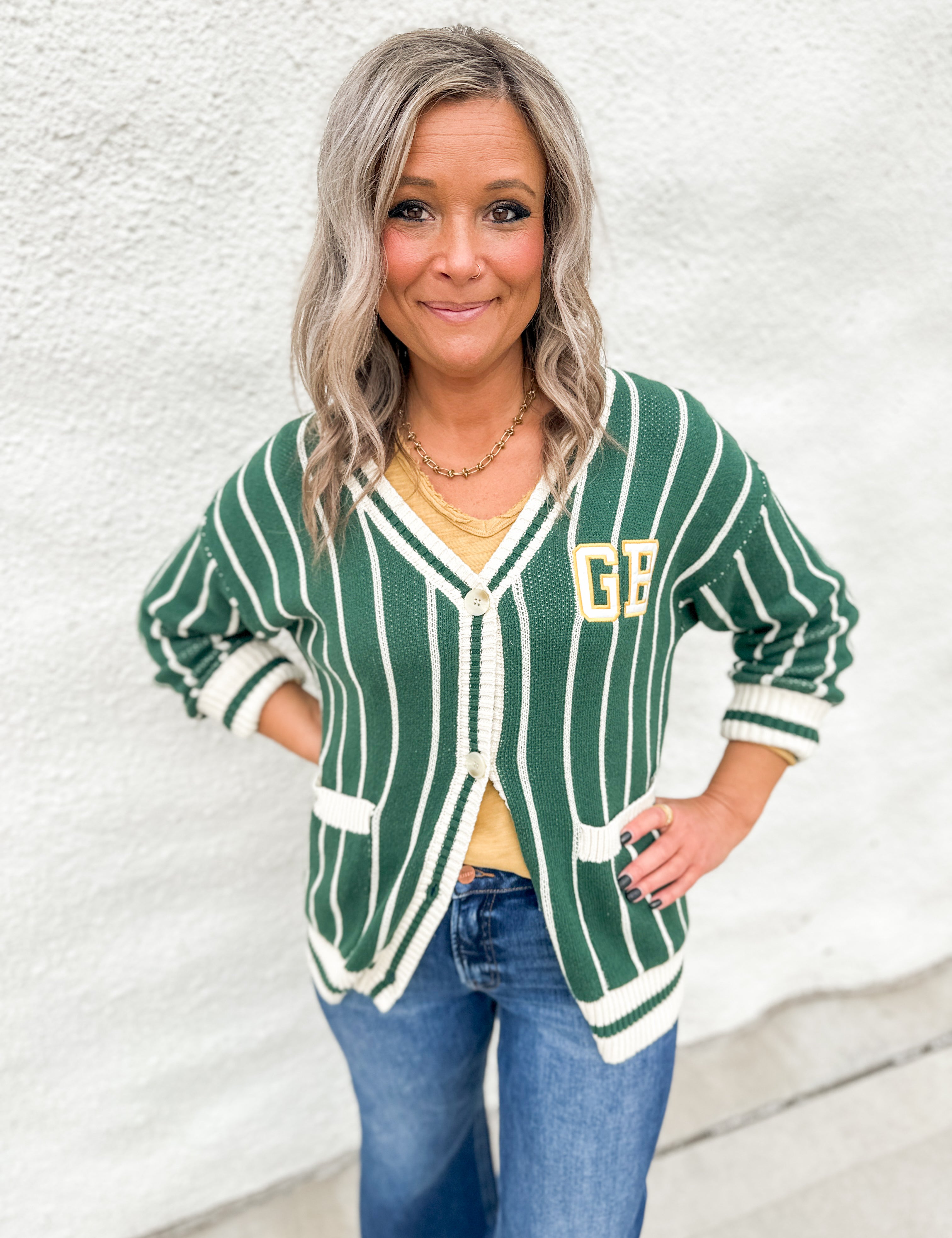 A girl wearing a green and white striped cardigan with GB on the front pocket area wearing blue jeans in front of a white wall.