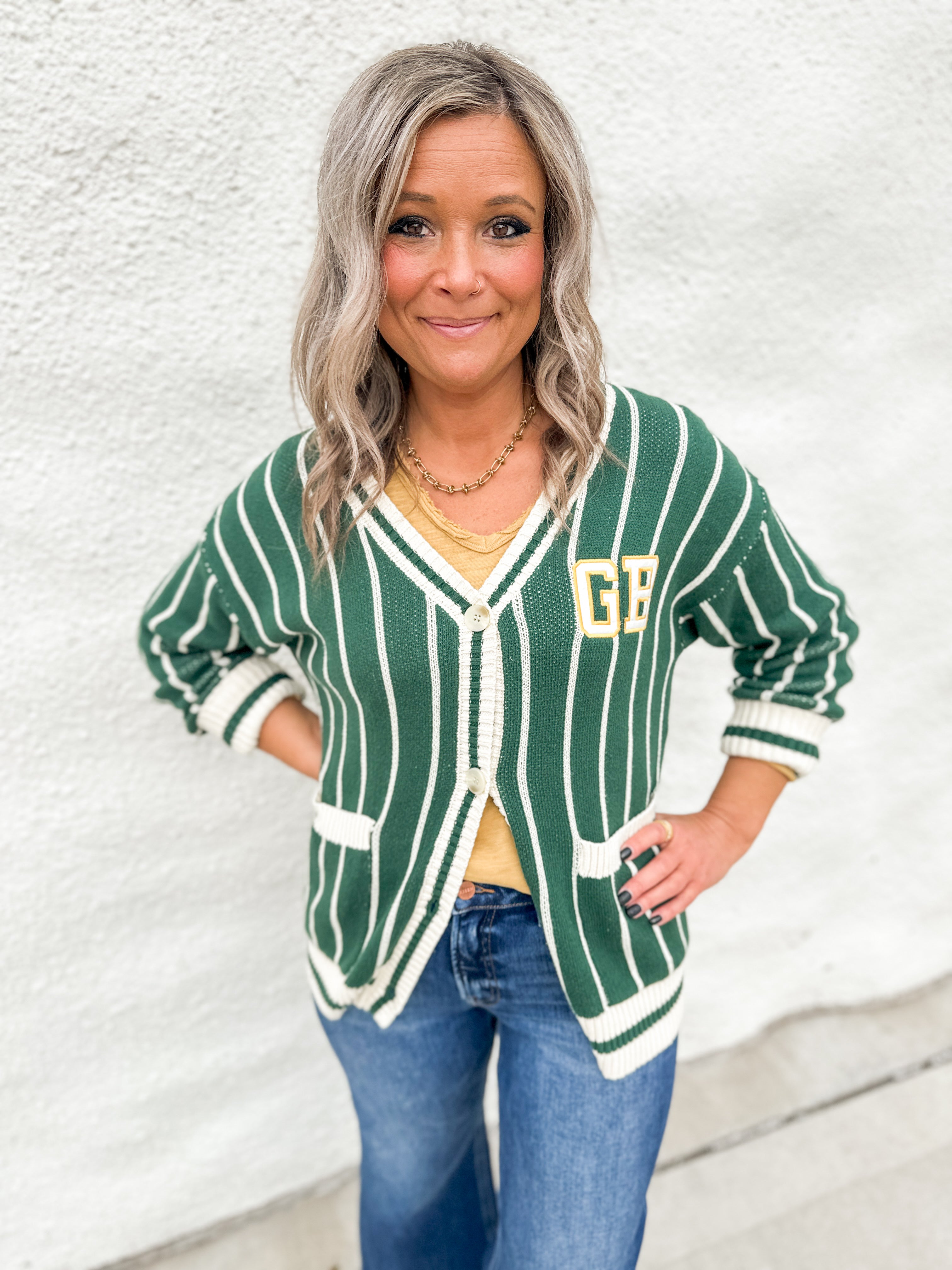 A girl wearing a green and white striped cardigan with GB on the front pocket area wearing blue jeans in front of a white wall.
