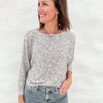 A woman wearing a textured, melange brown, cream & light gray knit sweater and gray-wash blue jeans, standing against a white stucco wall.