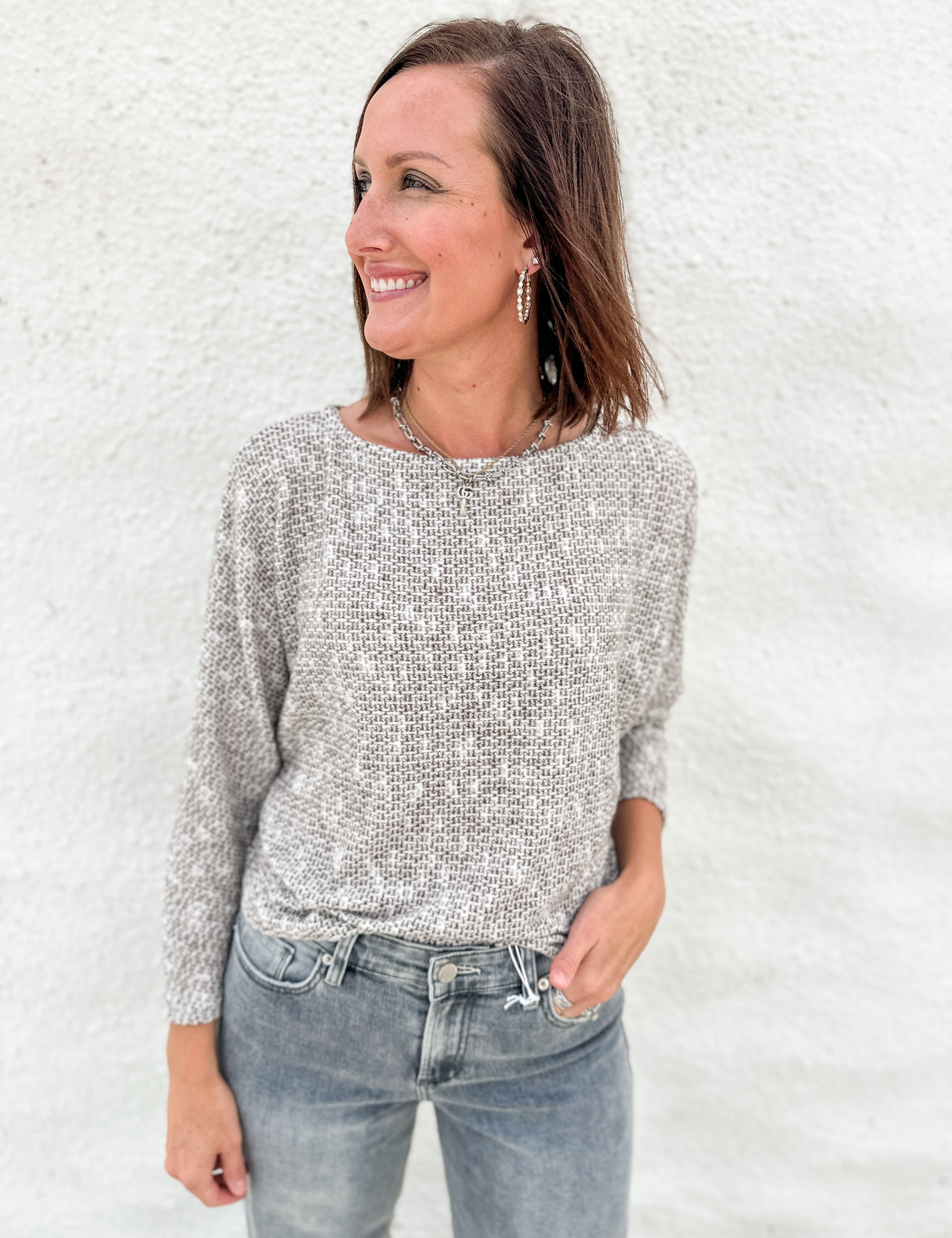 A woman wearing a textured, melange brown, cream & light gray knit sweater and gray-wash blue jeans, standing against a white stucco wall.