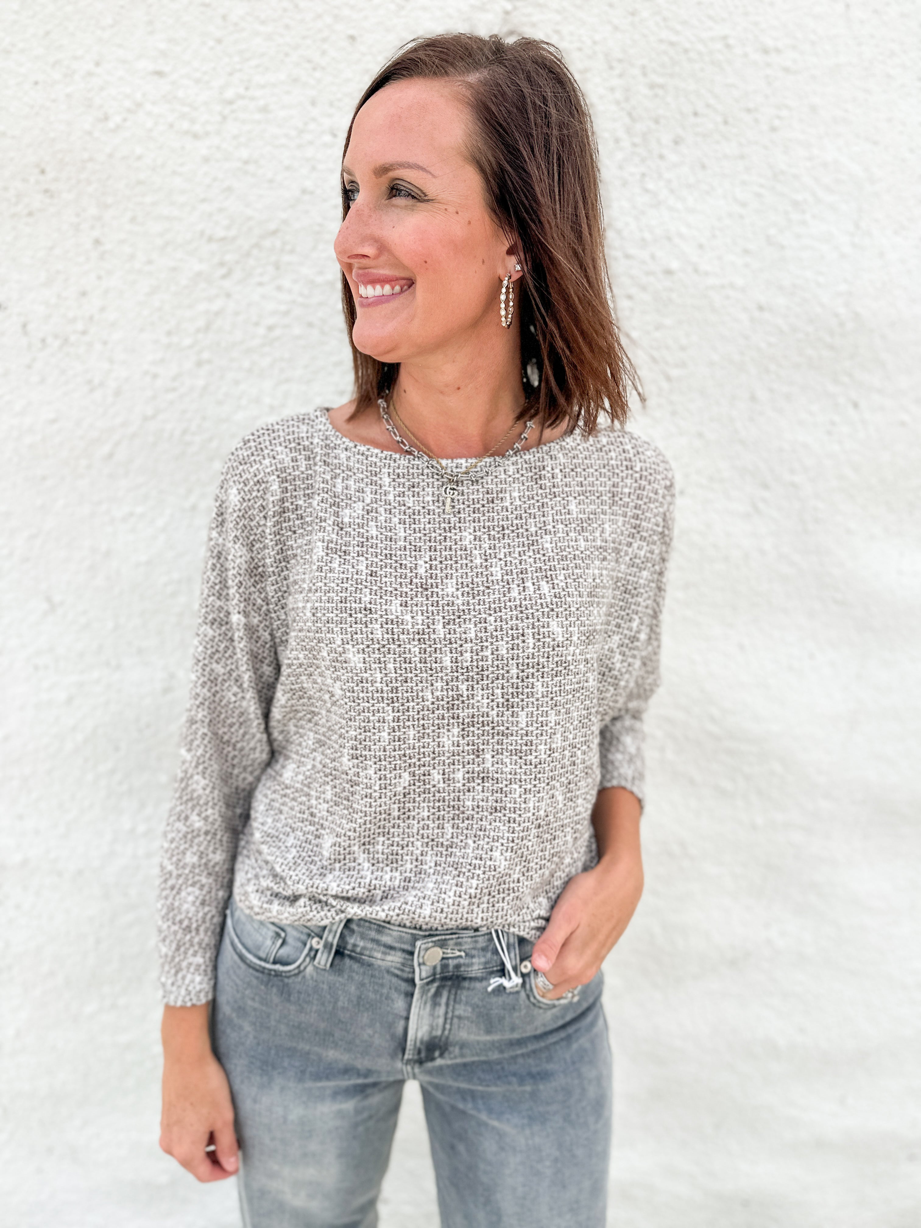 A woman wearing a textured, melange brown, cream & light gray knit sweater and gray-wash blue jeans, standing against a white stucco wall.
