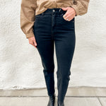 A person wearing black slim straight jeans, a tan button-up shirt, and black pointed-toe ankle boots, standing against a textured off-white wall.