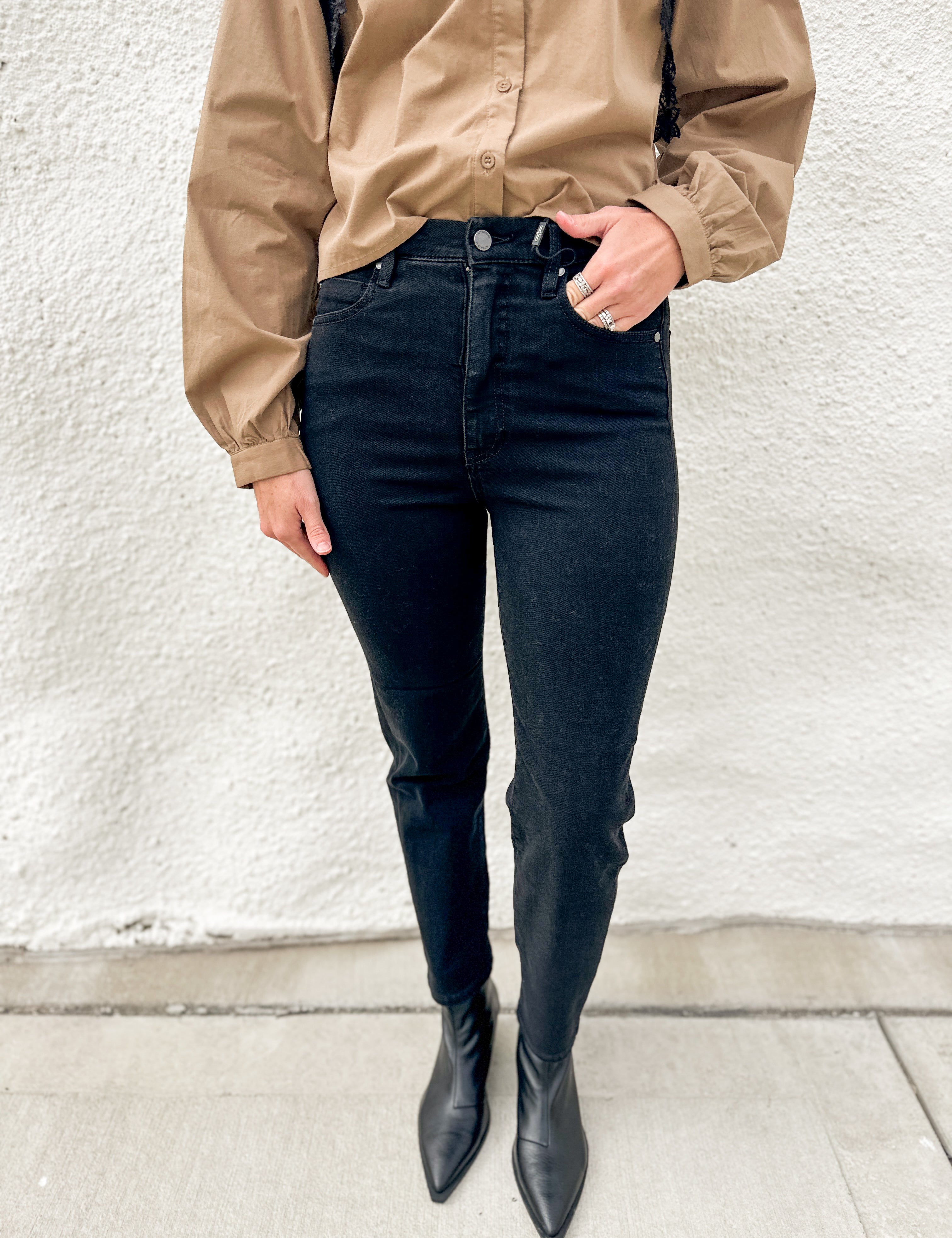 A person wearing black slim straight jeans, a tan button-up shirt, and black pointed-toe ankle boots, standing against a textured off-white wall.