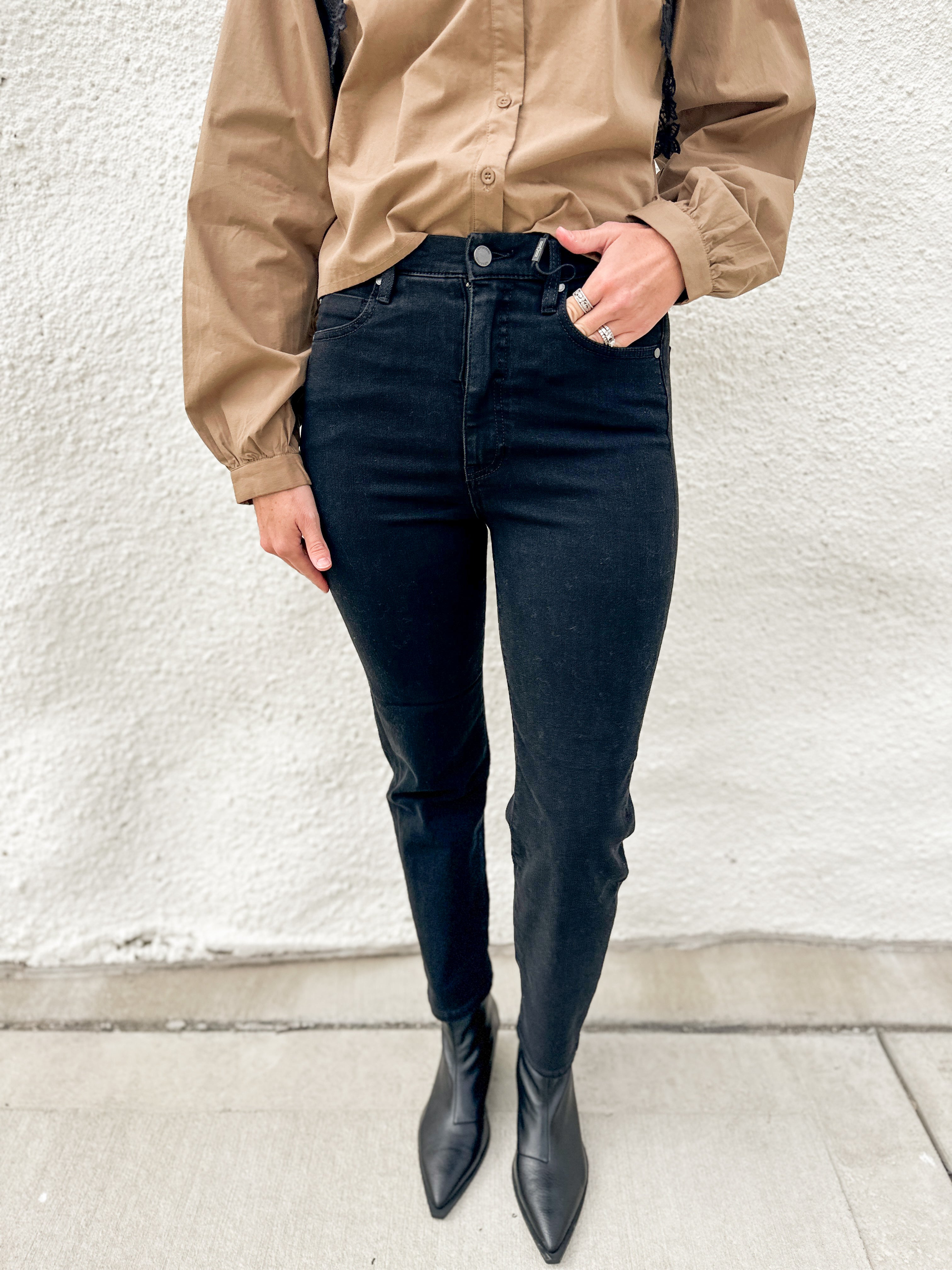 A person wearing black slim straight jeans, a tan button-up shirt, and black pointed-toe ankle boots, standing against a textured off-white wall.