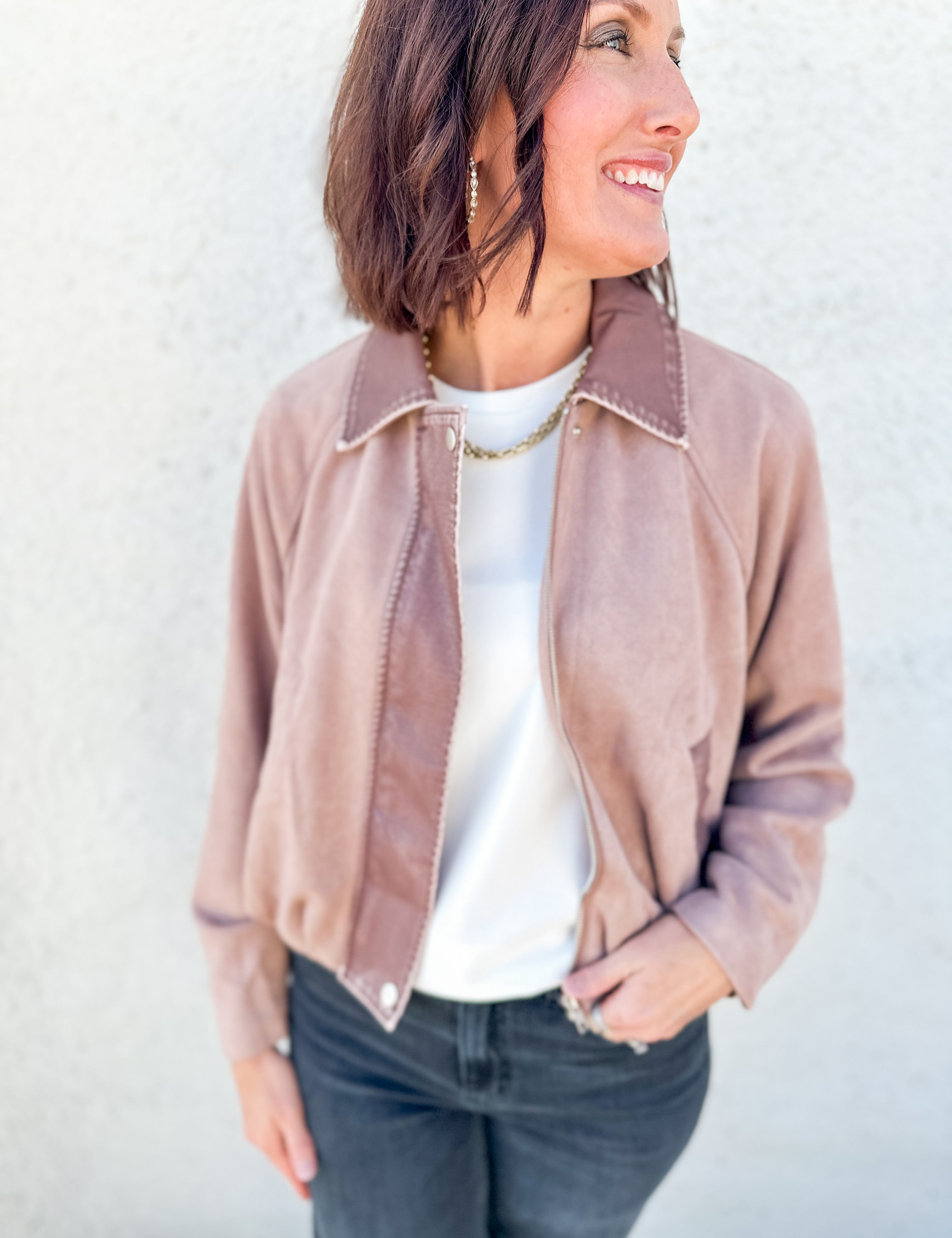 A woman wearing a dusty rose faux suede cropped bubble hem jacket over a white t-shirt and dark gray jeans, standing against a light textured wall.