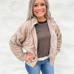 A smiling woman with gray hair stands against a white textured wall wearing a light beige quilted zip-up jacket over a brown top and blue jeans.