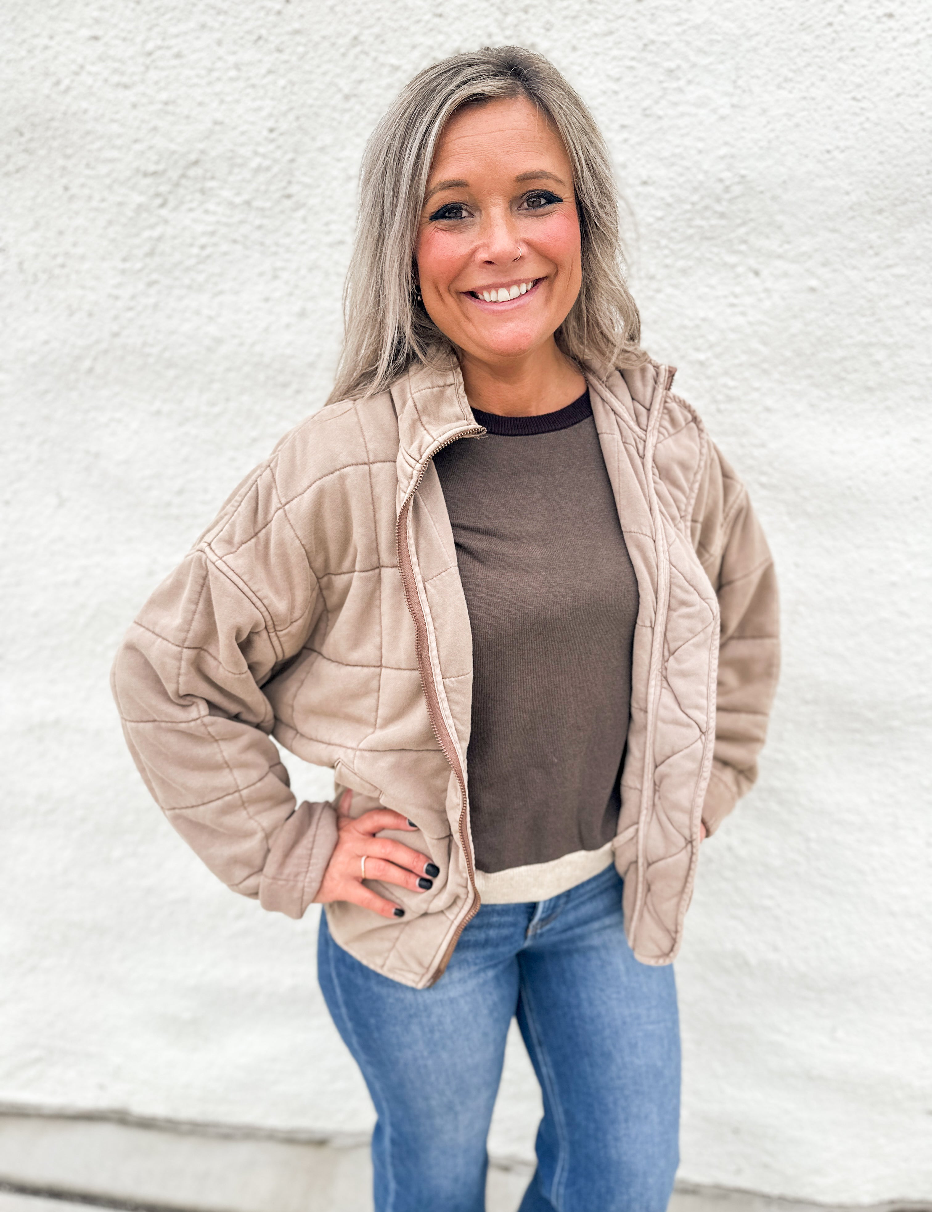 A smiling woman with gray hair stands against a white textured wall wearing a light beige quilted zip-up jacket over a brown top and blue jeans.