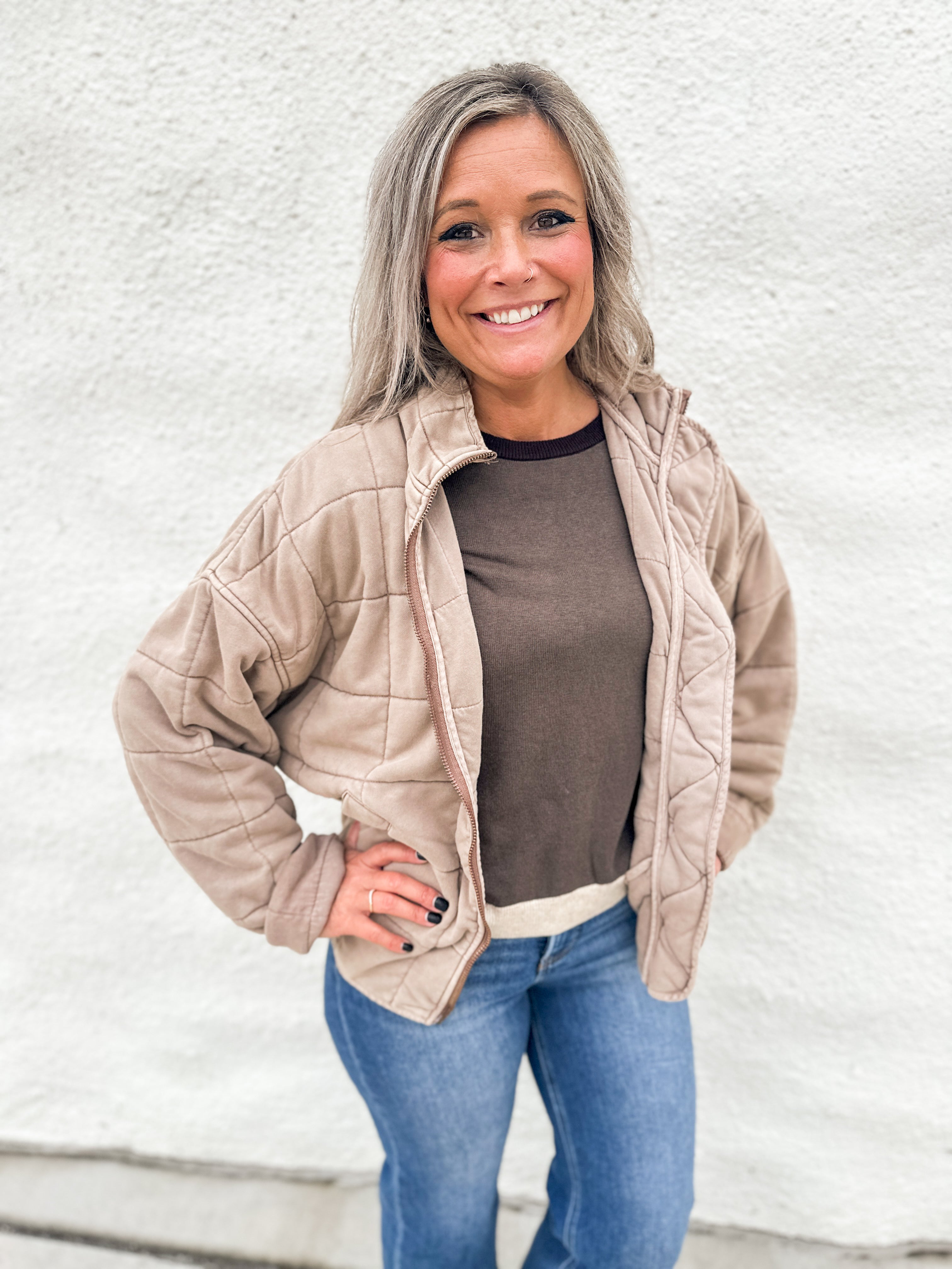 A smiling woman with gray hair stands against a white textured wall wearing a light beige quilted zip-up jacket over a brown top and blue jeans.