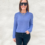A woman wearing a textured blue knit sweater from Free People, black sunglasses, and black pants stands against a white stucco wall.