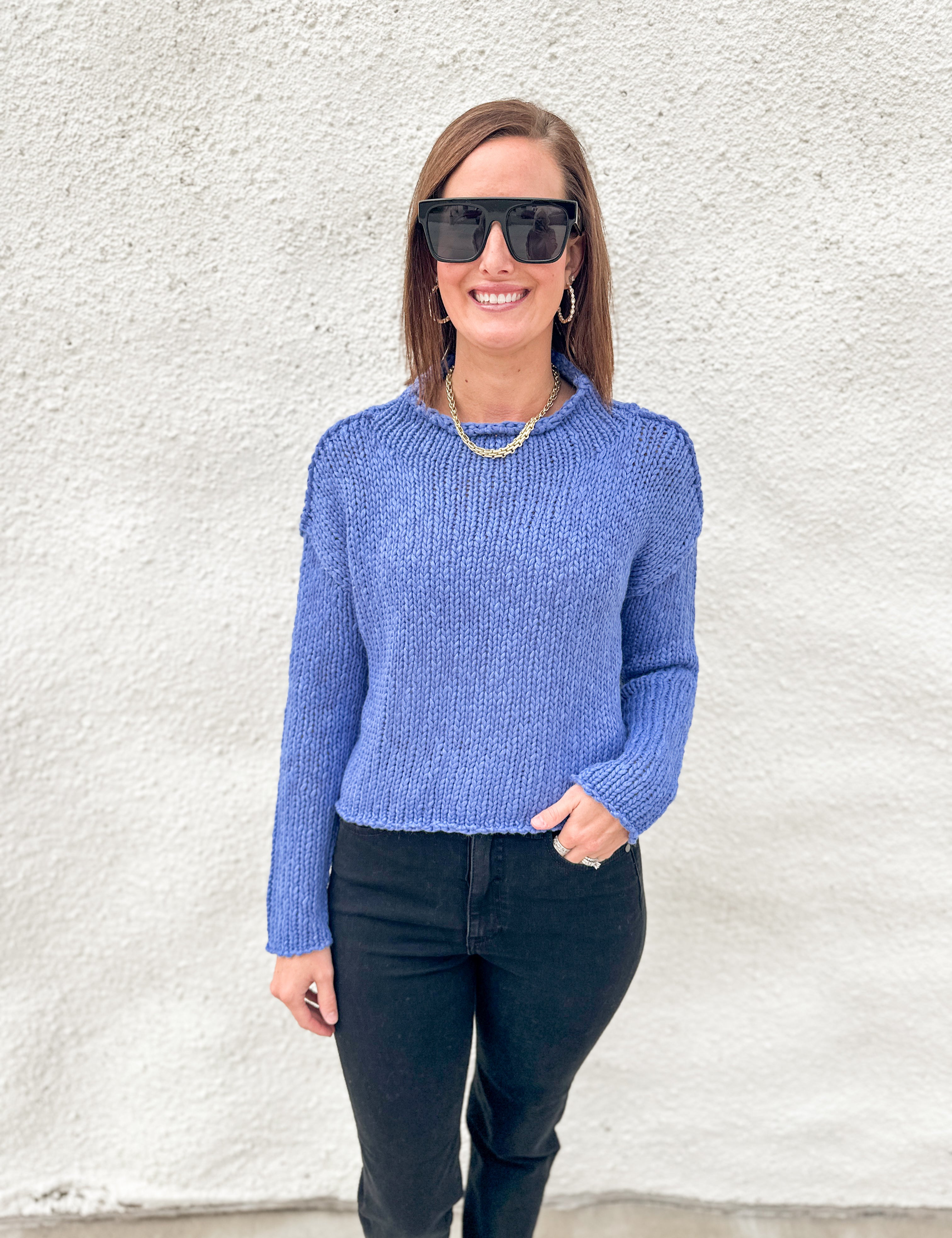 A woman wearing a textured blue knit sweater from Free People, black sunglasses, and black pants stands against a white stucco wall.