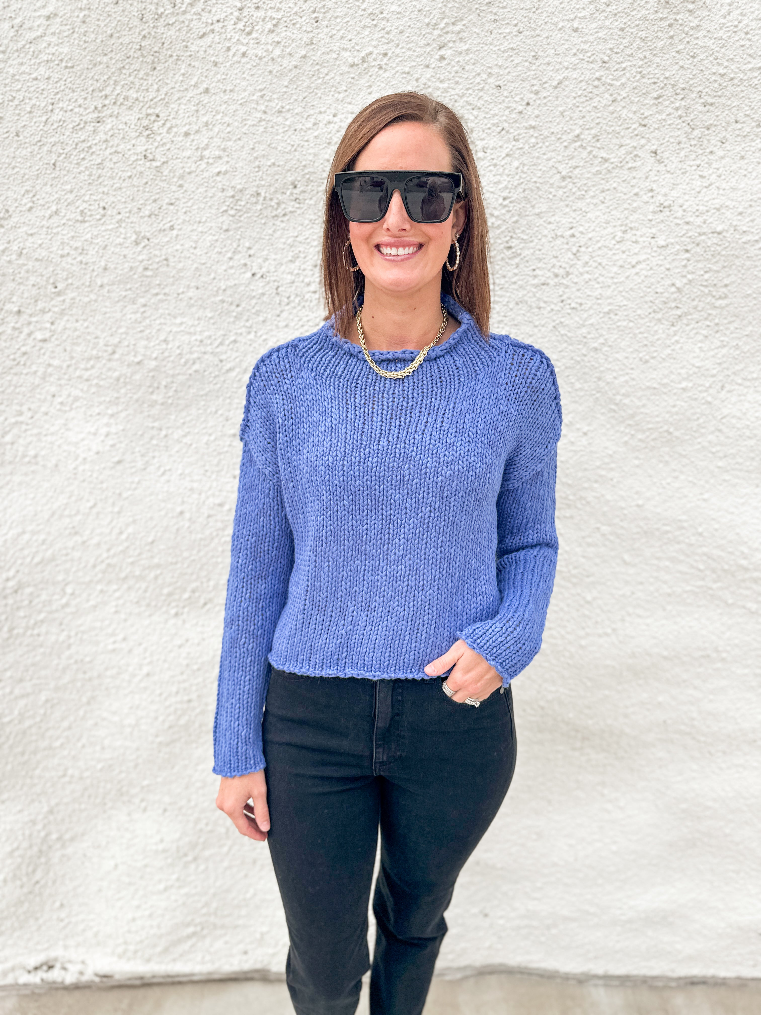 A woman wearing a textured blue knit sweater from Free People, black sunglasses, and black pants stands against a white stucco wall.