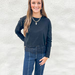 A woman modeling a black long-sleeve stretch crewneck top with lace detail at bottom hem and dark blue jeans, standing against a white textured wall.
