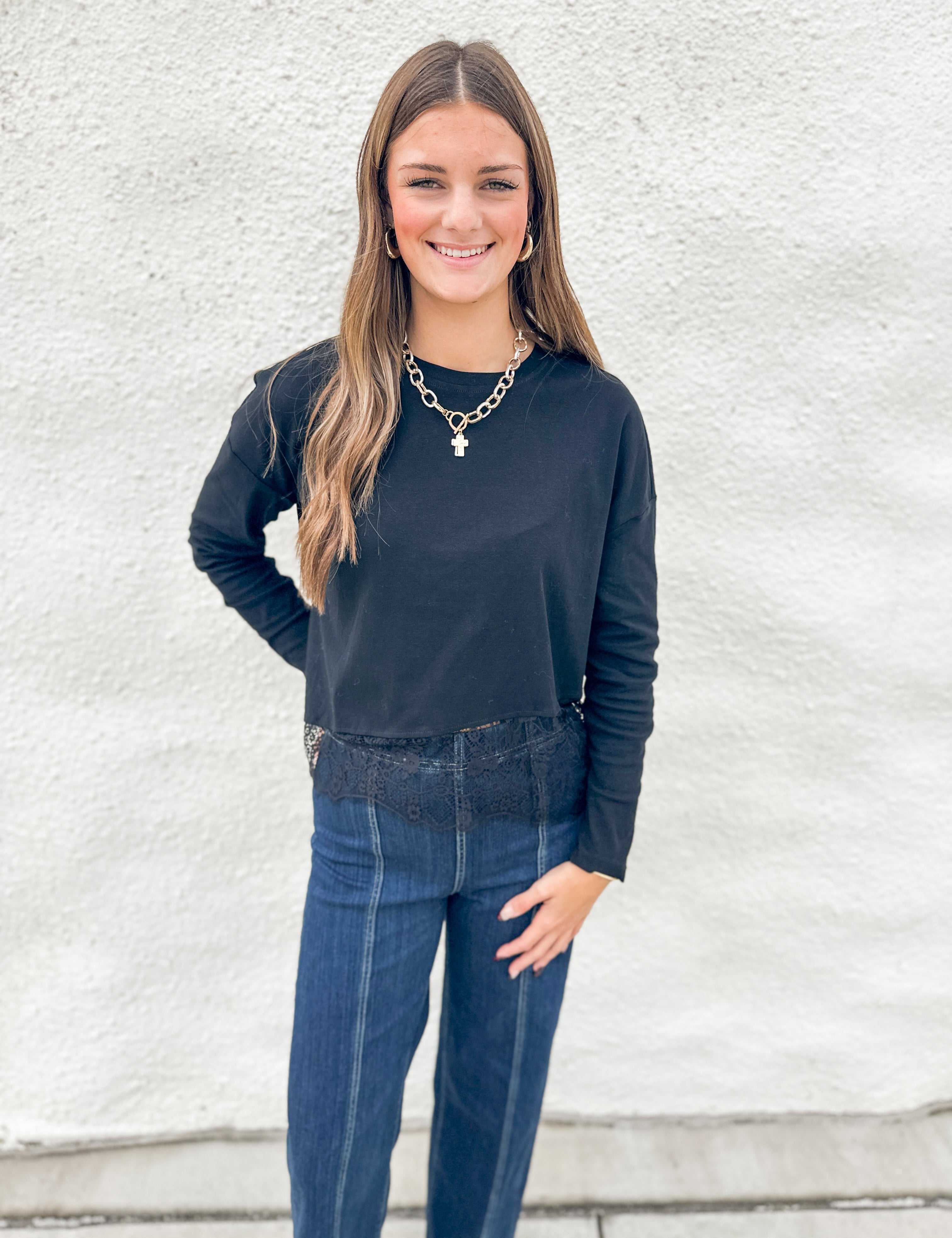 A woman modeling a black long-sleeve stretch crewneck top with lace detail at bottom hem and dark blue jeans, standing against a white textured wall.