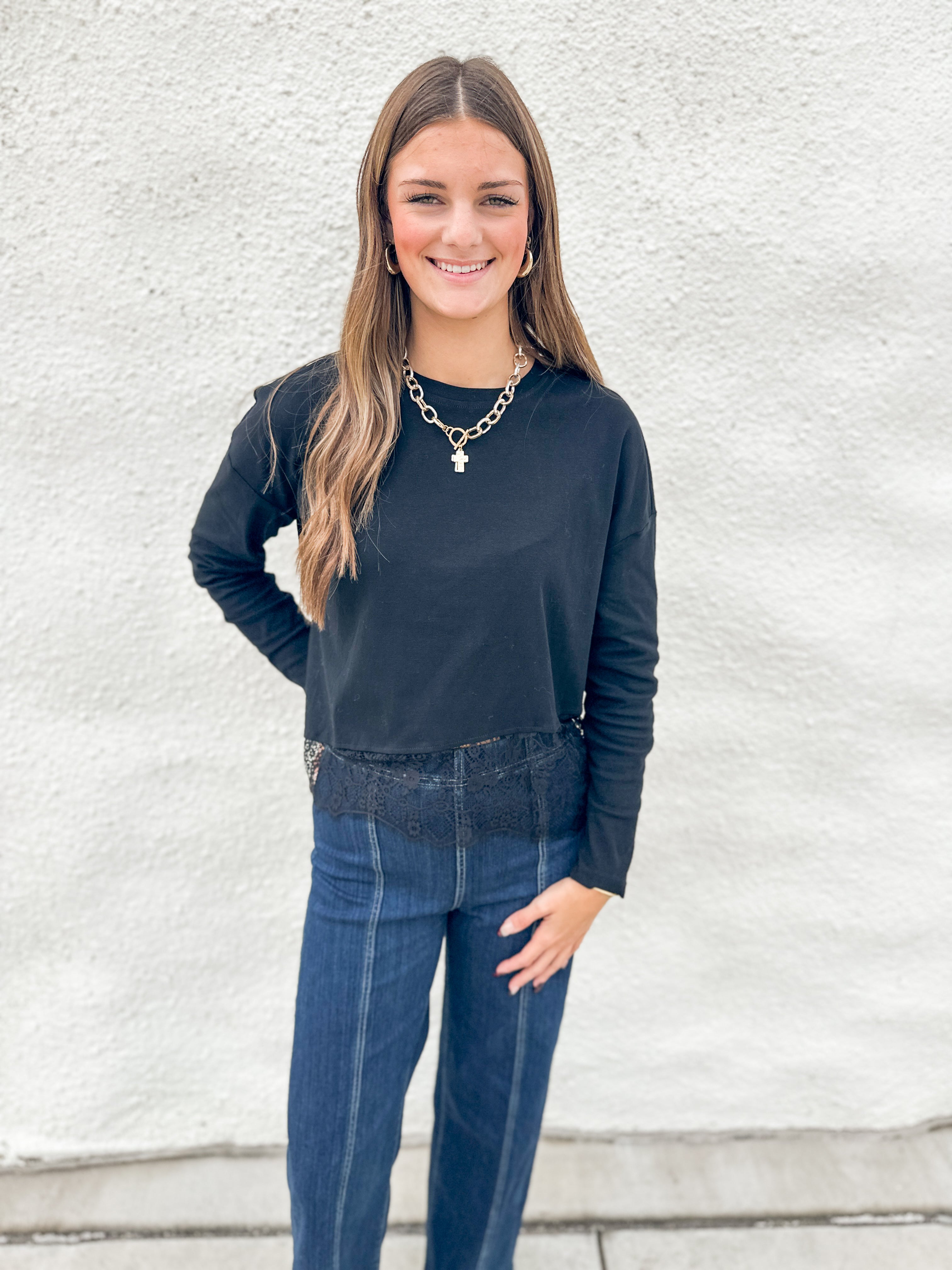 A woman modeling a black long-sleeve stretch crewneck top with lace detail at bottom hem and dark blue jeans, standing against a white textured wall.