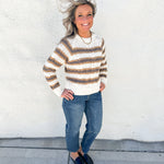 A woman stands outdoors against a white stucco wall wearing a cream and brown horizontally striped knit sweater, blue jeans, and dark loafers.