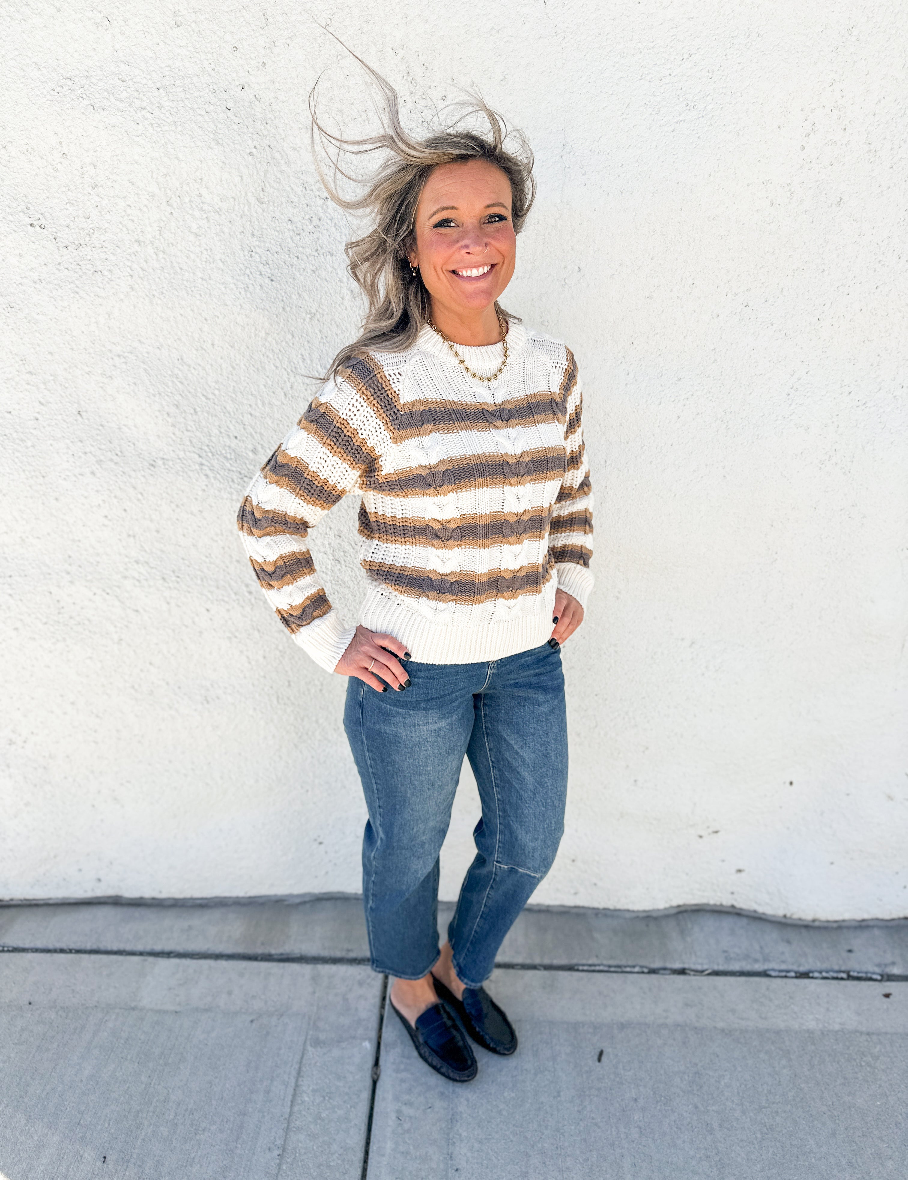 A woman stands outdoors against a white stucco wall wearing a cream and brown horizontally striped knit sweater, blue jeans, and dark loafers.