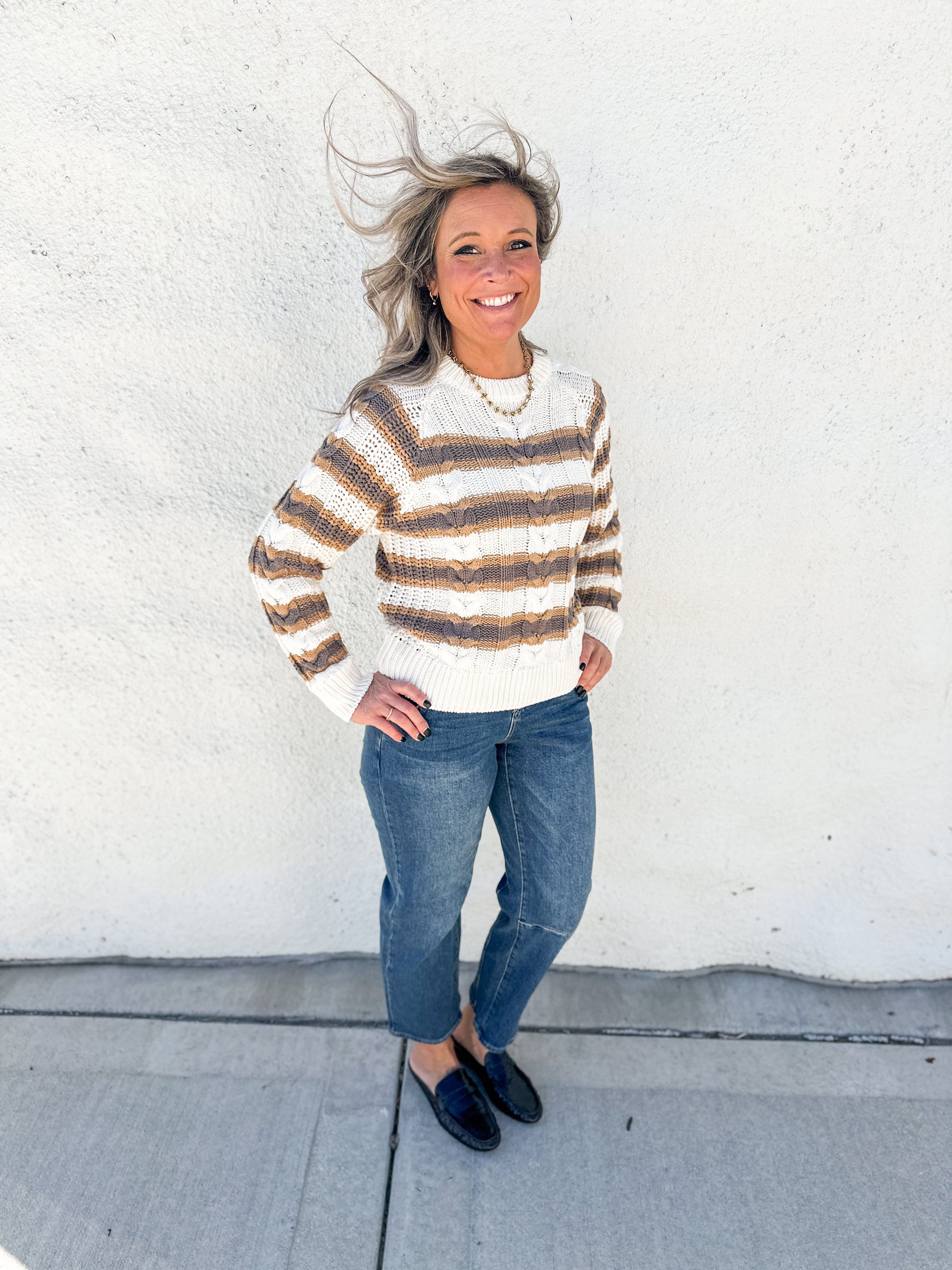 A woman stands outdoors against a white stucco wall wearing a cream and brown horizontally striped knit sweater, blue jeans, and dark loafers.