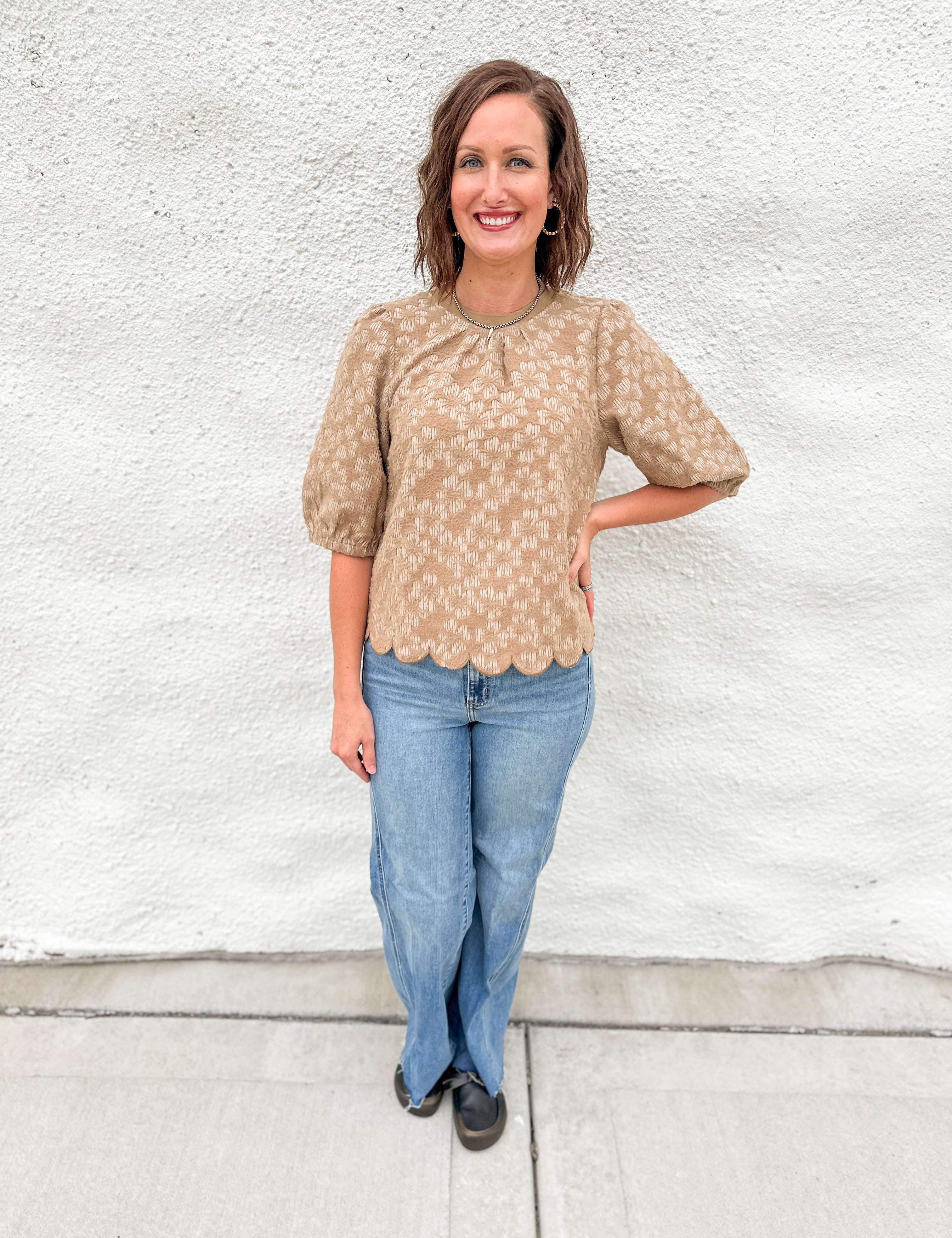 A woman wearing a tam floral 3/4 sleeve top with scalloped hem and light denim jeans with black mules in front of a white wall.