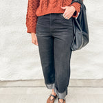 A person wearing a rust-orange cable-knit sweater, dark charcoal gray cuffed jeans, and brown Mary Jane flats, carrying a black leather shoulder bag against a white stucco wall.