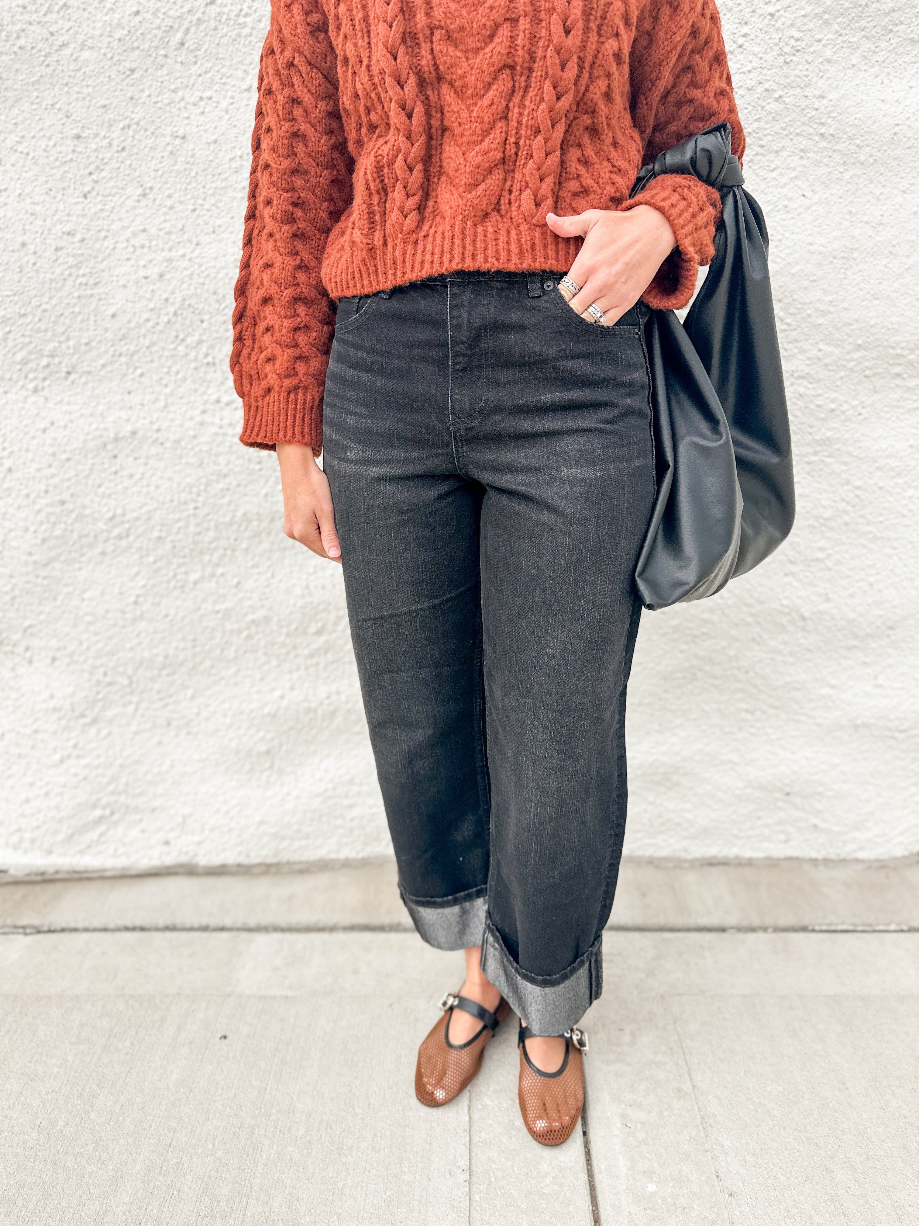 A person wearing a rust-orange cable-knit sweater, dark charcoal gray cuffed jeans, and brown Mary Jane flats, carrying a black leather shoulder bag against a white stucco wall.